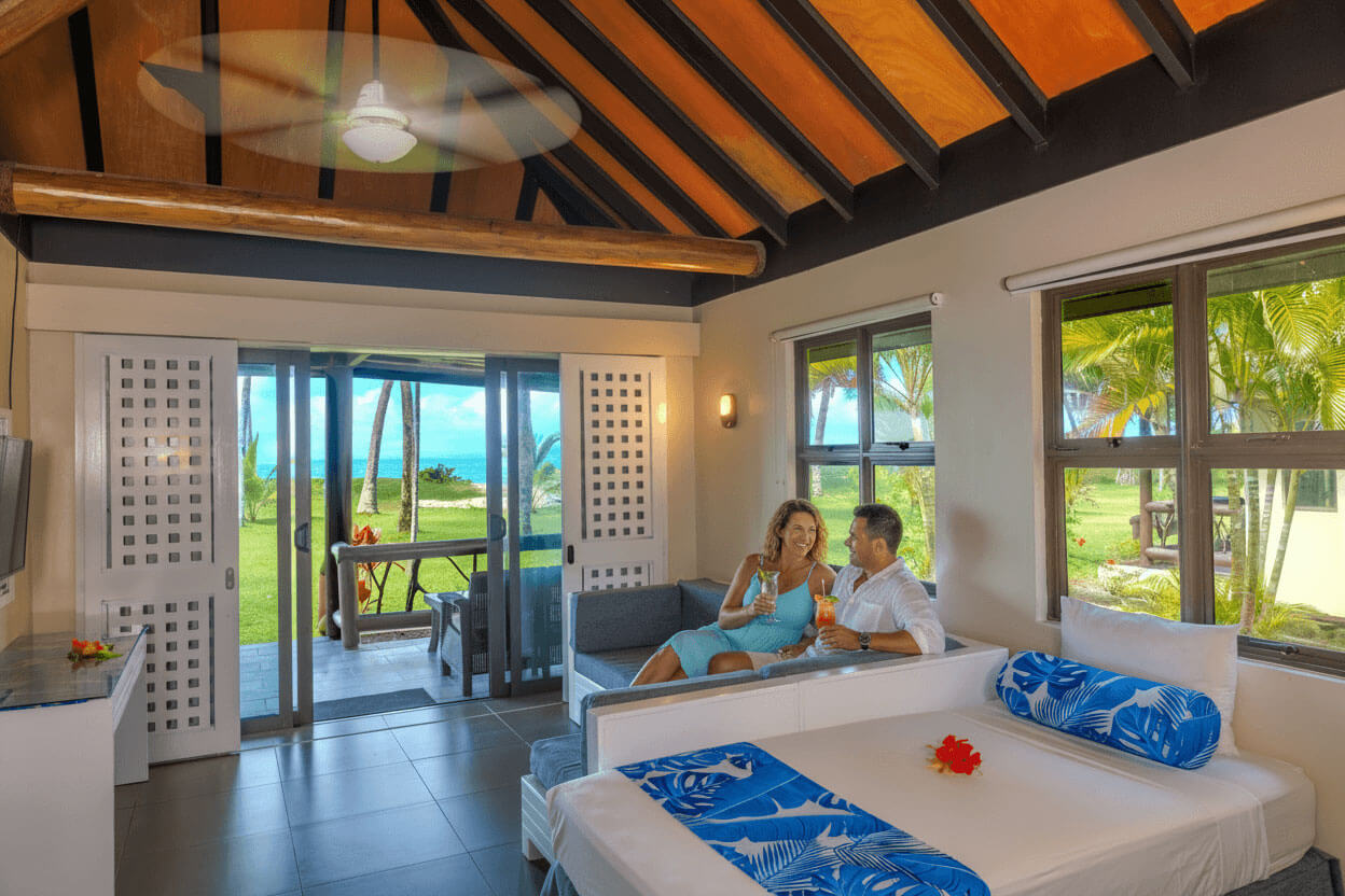 Couple enjoys a cocktail on the couch in a beachfront bure at Uprising Beach Resort, Fiji, with ocean views.