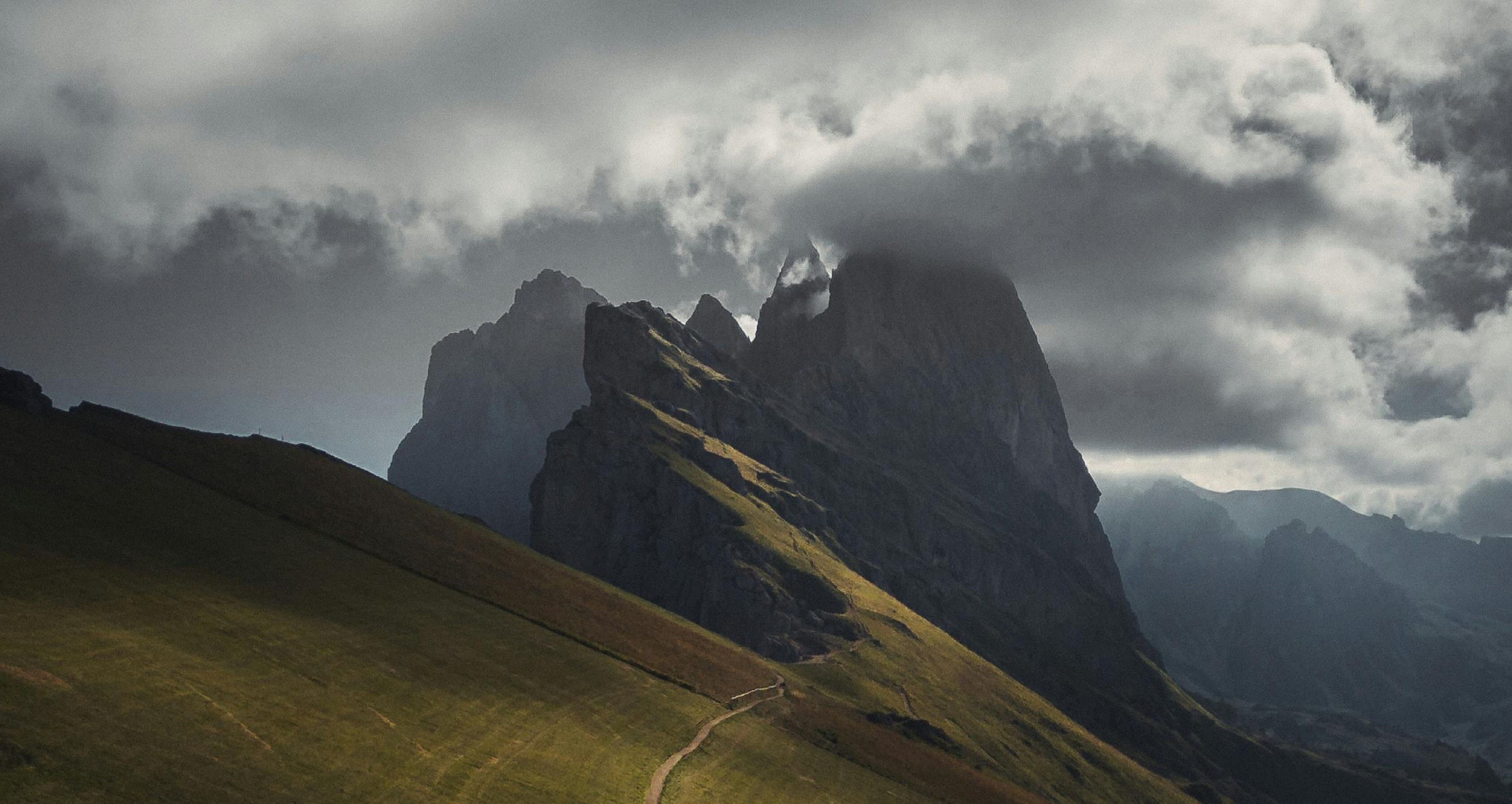 Winding footpath across a sloping green hillside leading toward jagged, dark rocky peaks partially shrouded in low clouds under a dramatic, moody sky.