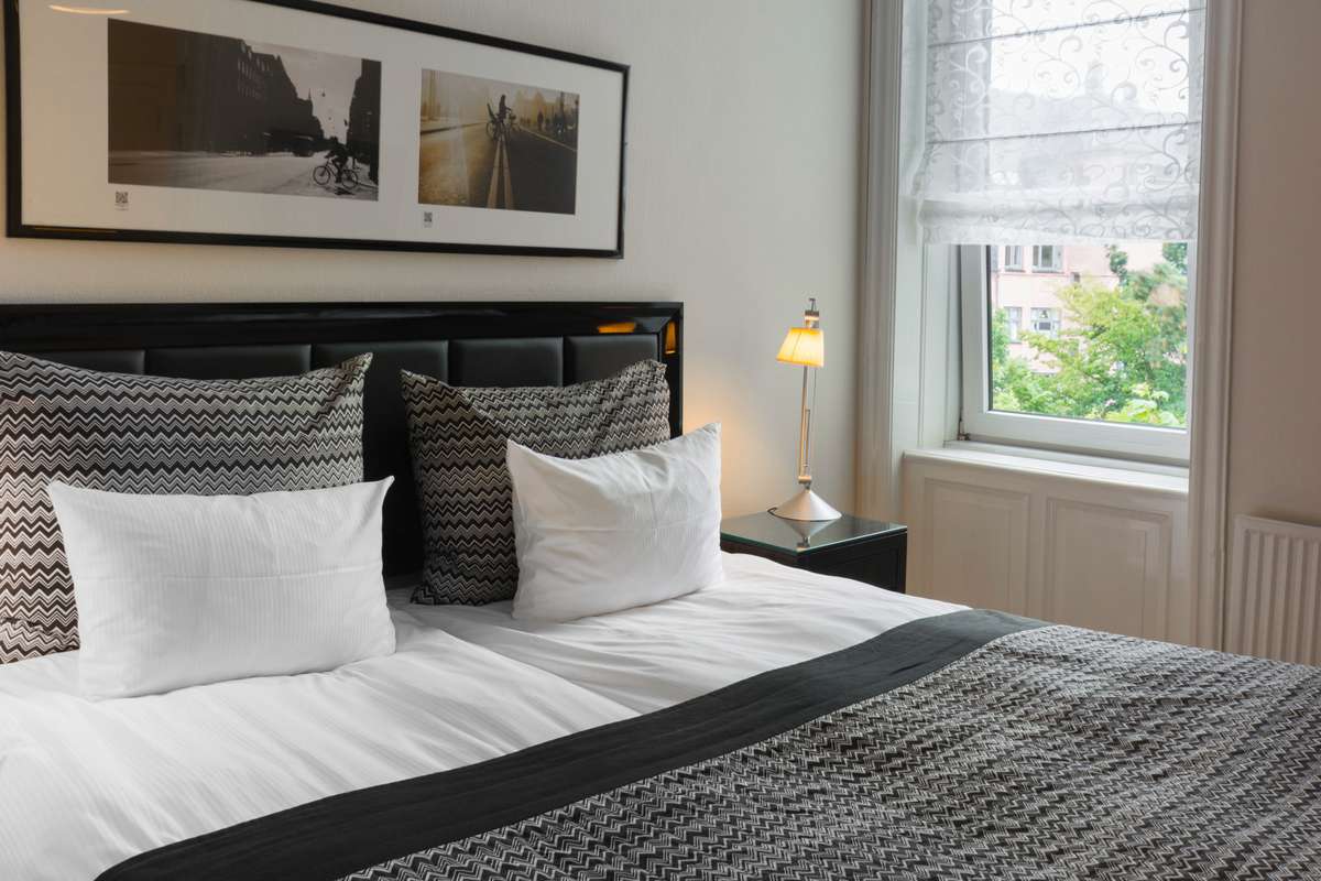 A hotel bed with white and grey patterned pillows sits before a large framed landscape photo and a window view of trees.