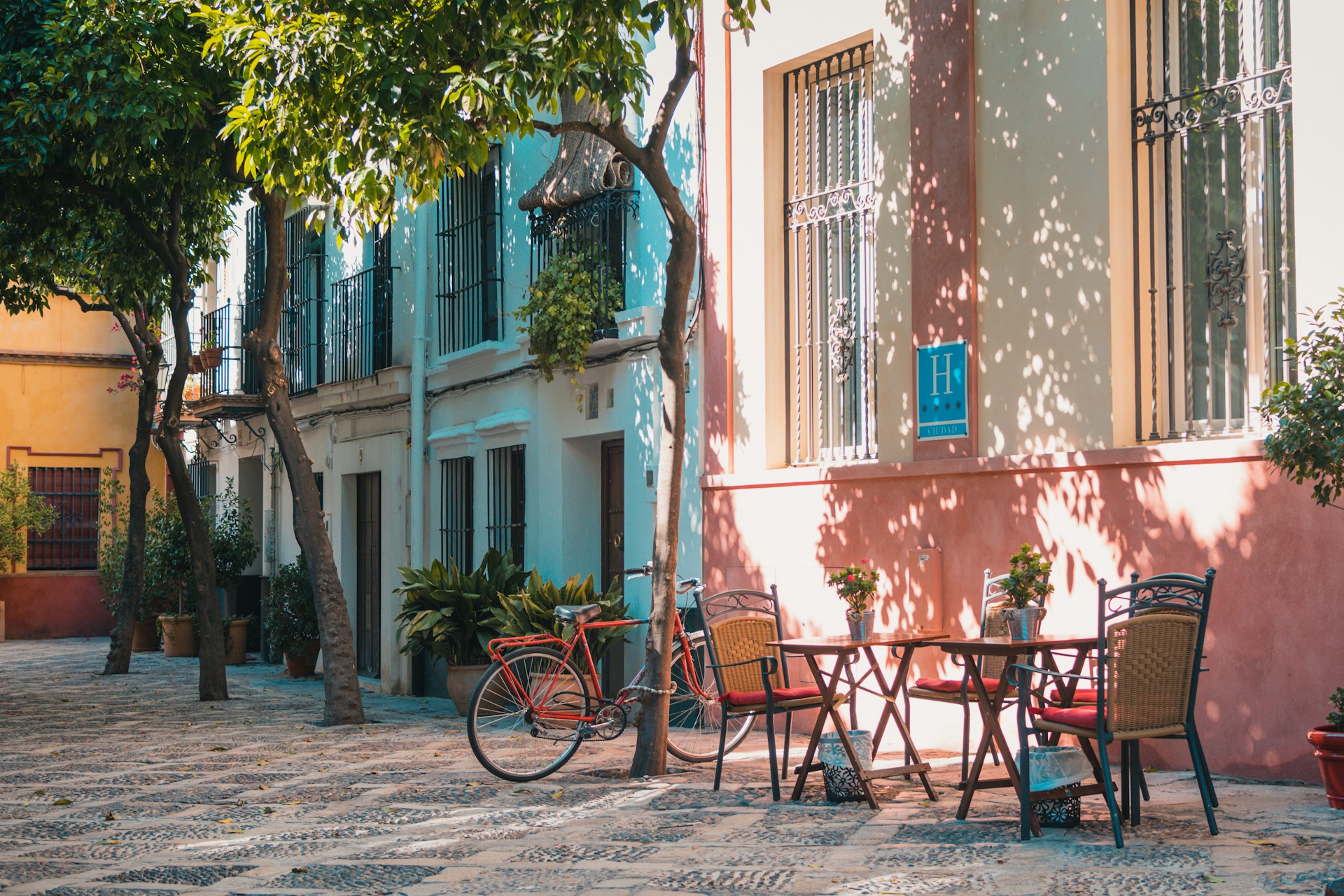 An outdoor scene of a quaint European cobblestone square or patio lined with trees and potted plants. On the right, a table with four chairs sits outside a building with faded peach and cream-colored walls. In the background, there are multi-story buildings with light blue-green facades and wrought iron window grates. A vintage red bicycle is parked near the tables.