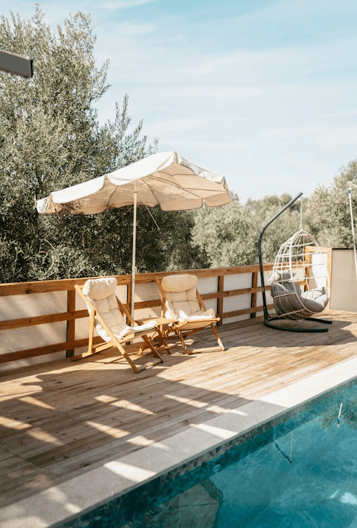 Shaded outdoor lounge area with beige umbrella, trees, and poolside on a tiled deck.
