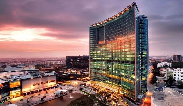 A green suspension bridge spanning across a body of water, with forested hills and residential homes in the background.