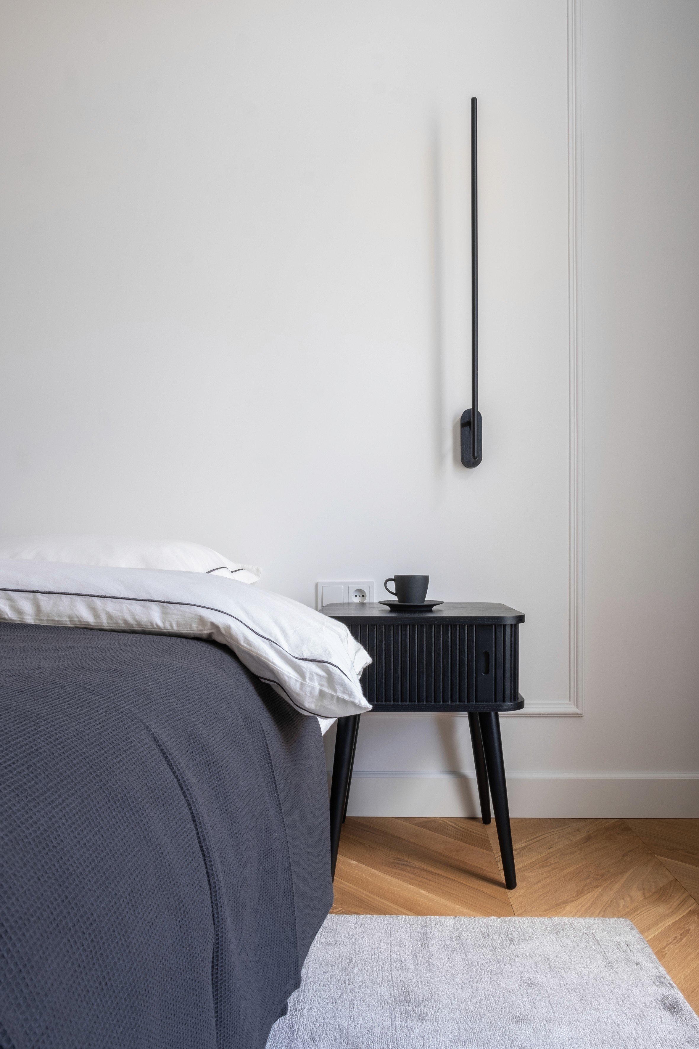 Minimalist bedroom with dark grey linen bedding, black ribbed cylindrical nightstand, and modern wall-mounted reading sconce
