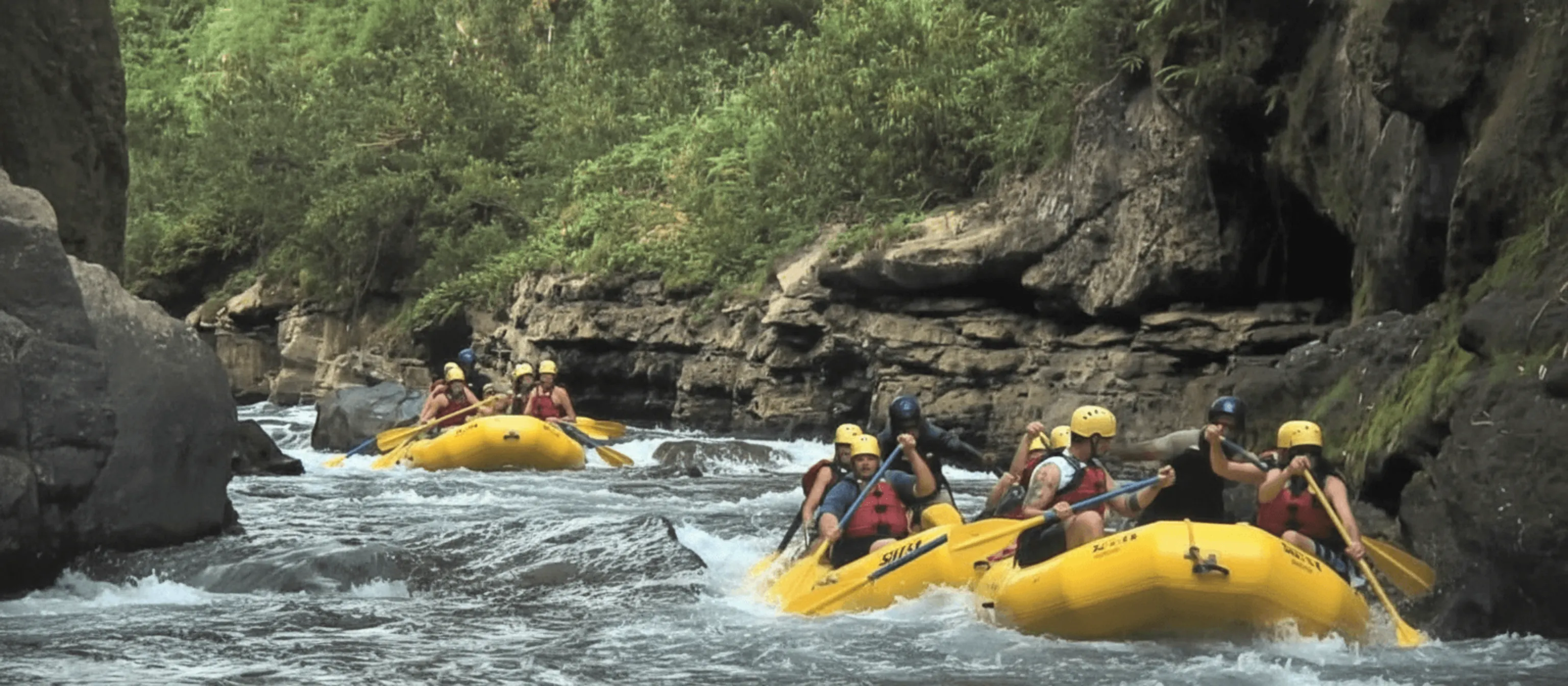 People in a yellow raft enjoy Pacific Harbour activities, cheering and paddling between jungle and rock walls.