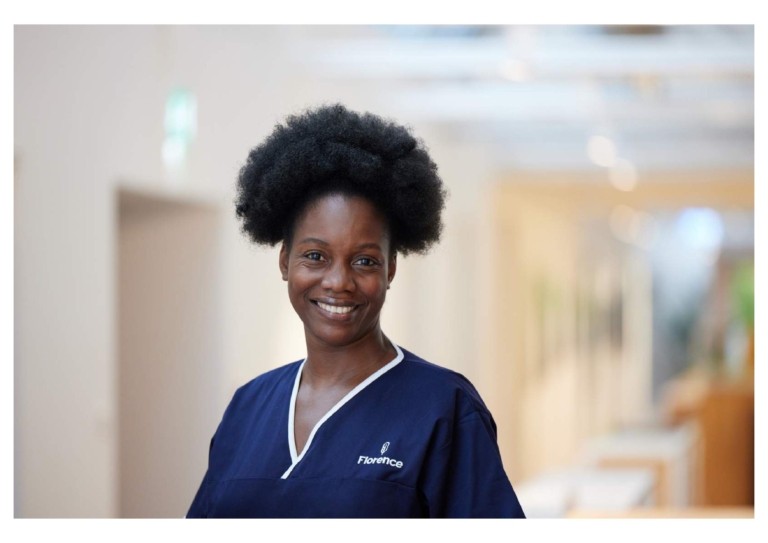 A female healthcare worker in a blue uniform standing in a bright healthcare corridor, representing nurses working long shifts