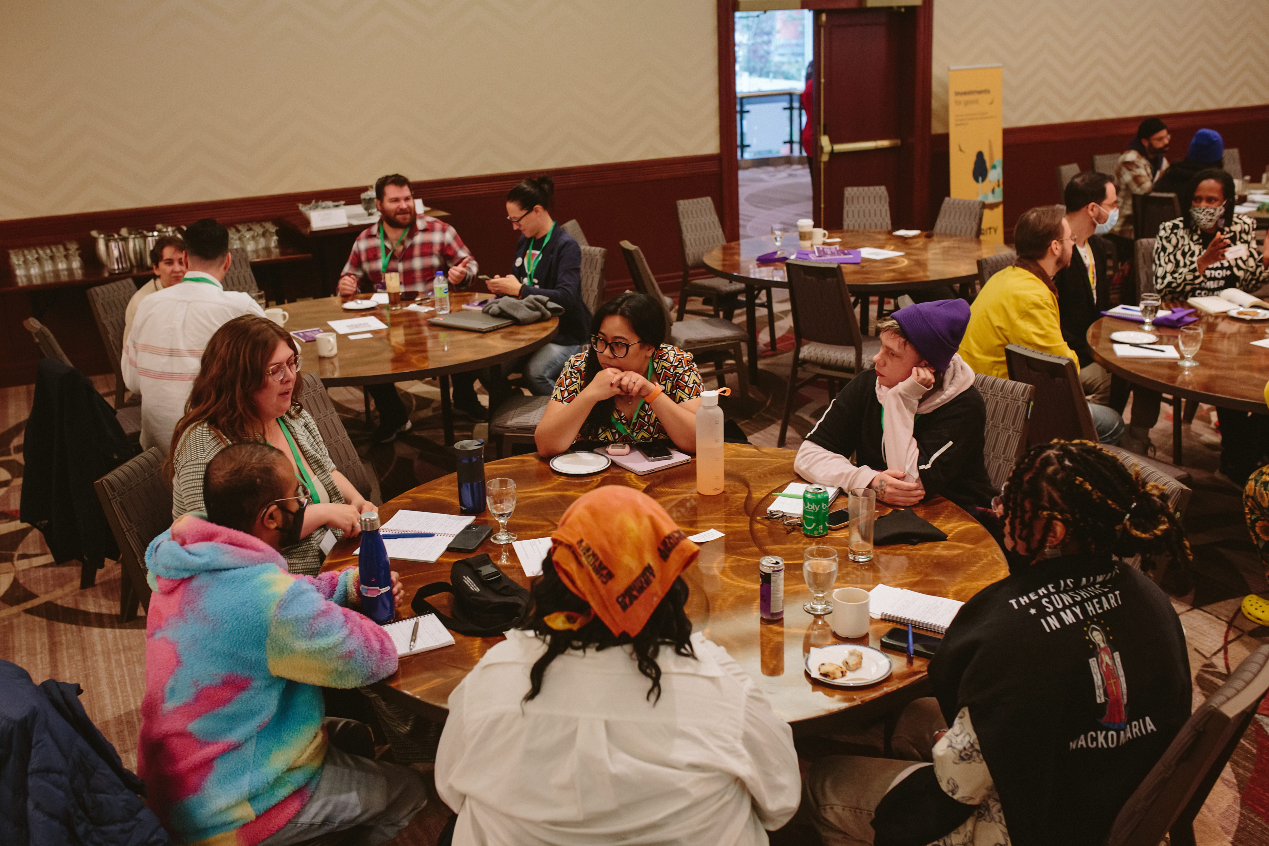 A diverse group of people gathered around tables, engaging in discussion in a well-lit room.