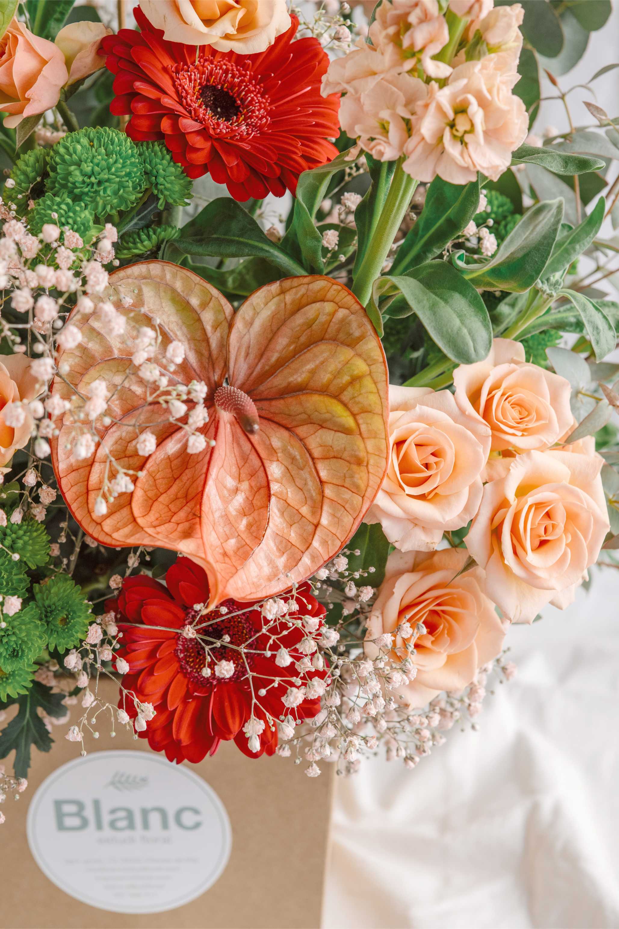 Pla detallat d’un arranjament floral amb anthurium ataronjat, gerbera vermella, roses préssec i gipsofila blanca, destacant la textura i combinació de colors suaus i intensos.