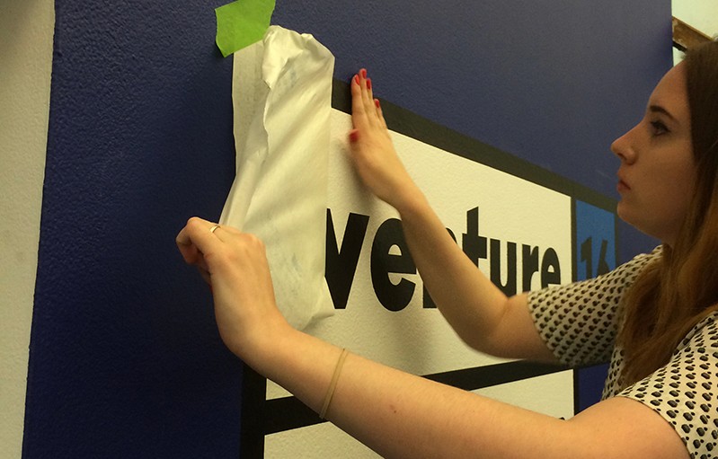 A woman sticking a venture 16 logo on a blue wall.