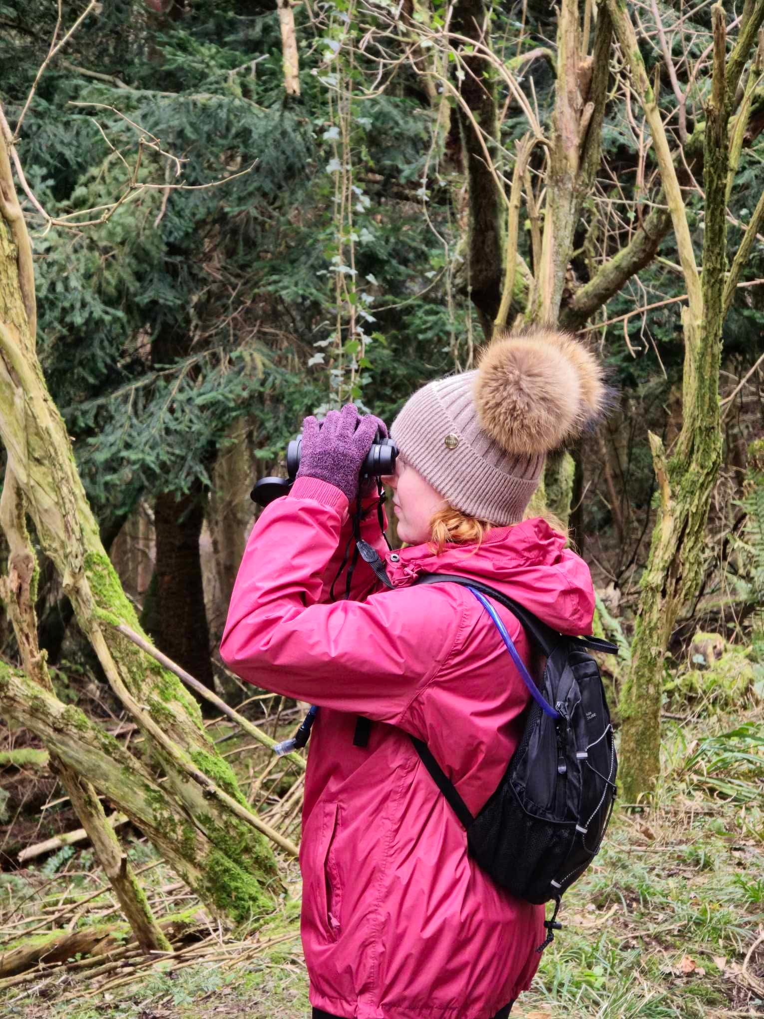 Eve wearing a bobblehat and waterproof coat surrounded by trees holding up binoculars to her face.