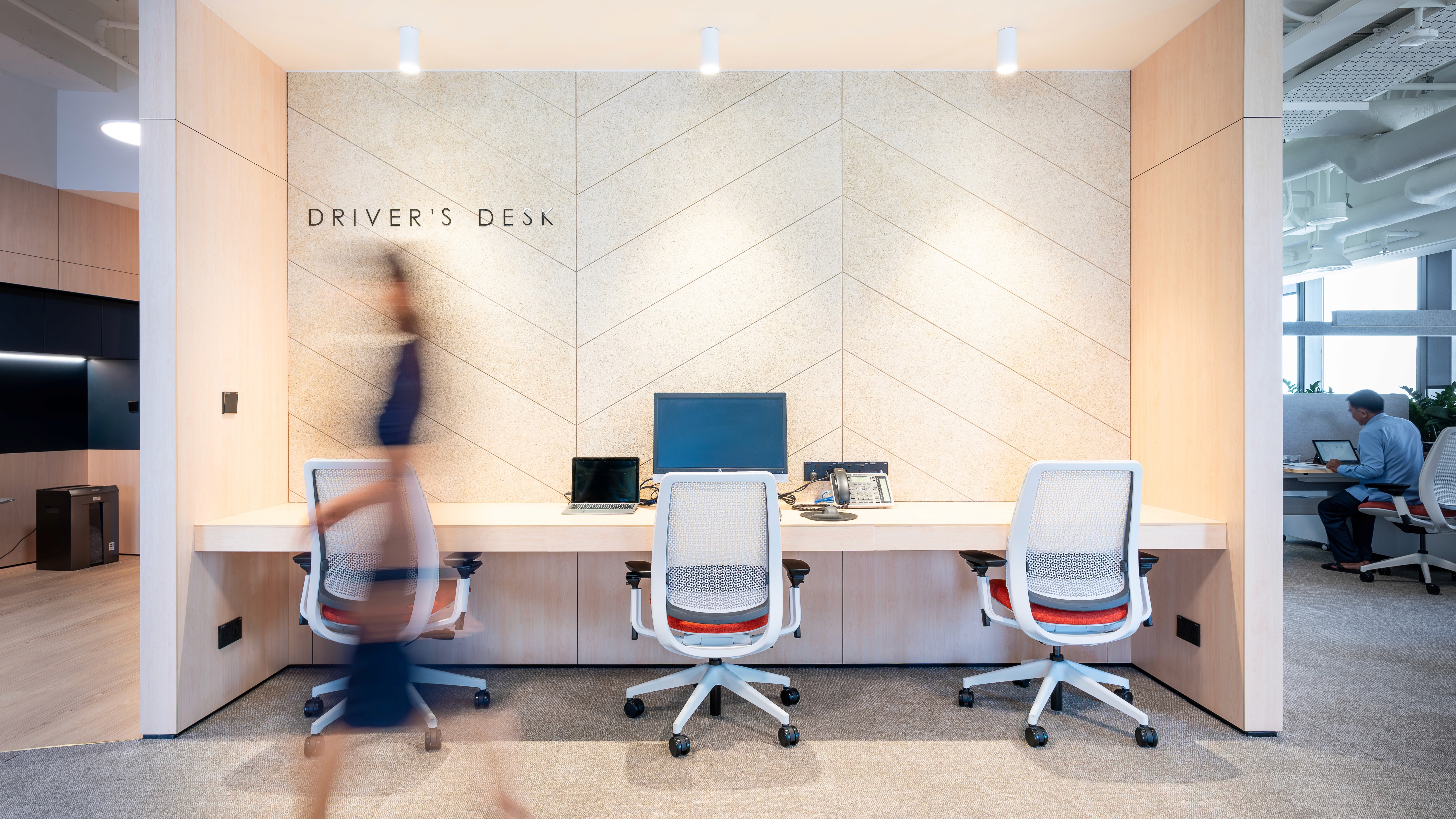 A modern office workstation area, featuring individual desks set against light-toned wooden walls with a subtle pattern, equipped with computers and ergonomic chairs.