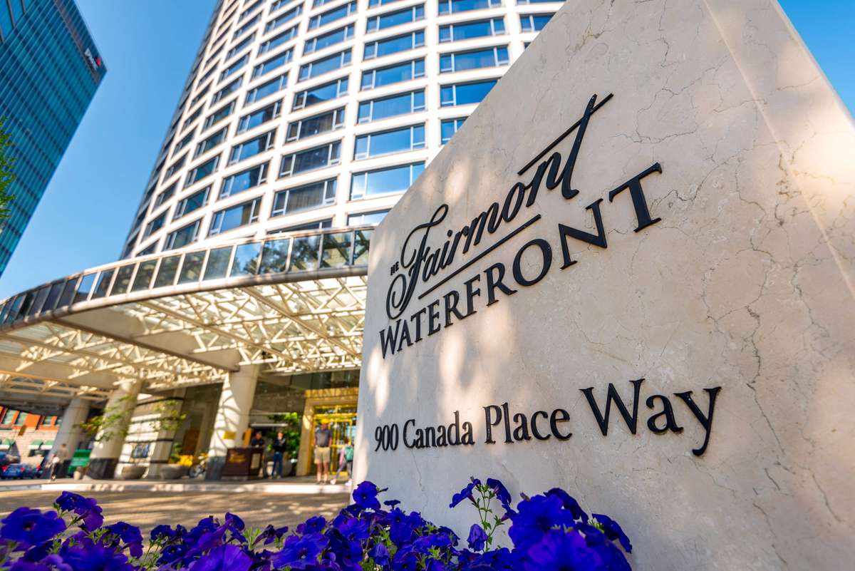 A stone wall with "Fairmont Waterfront" and "90 Canada Place Way" engraved in black lettering next to a modern hotel.