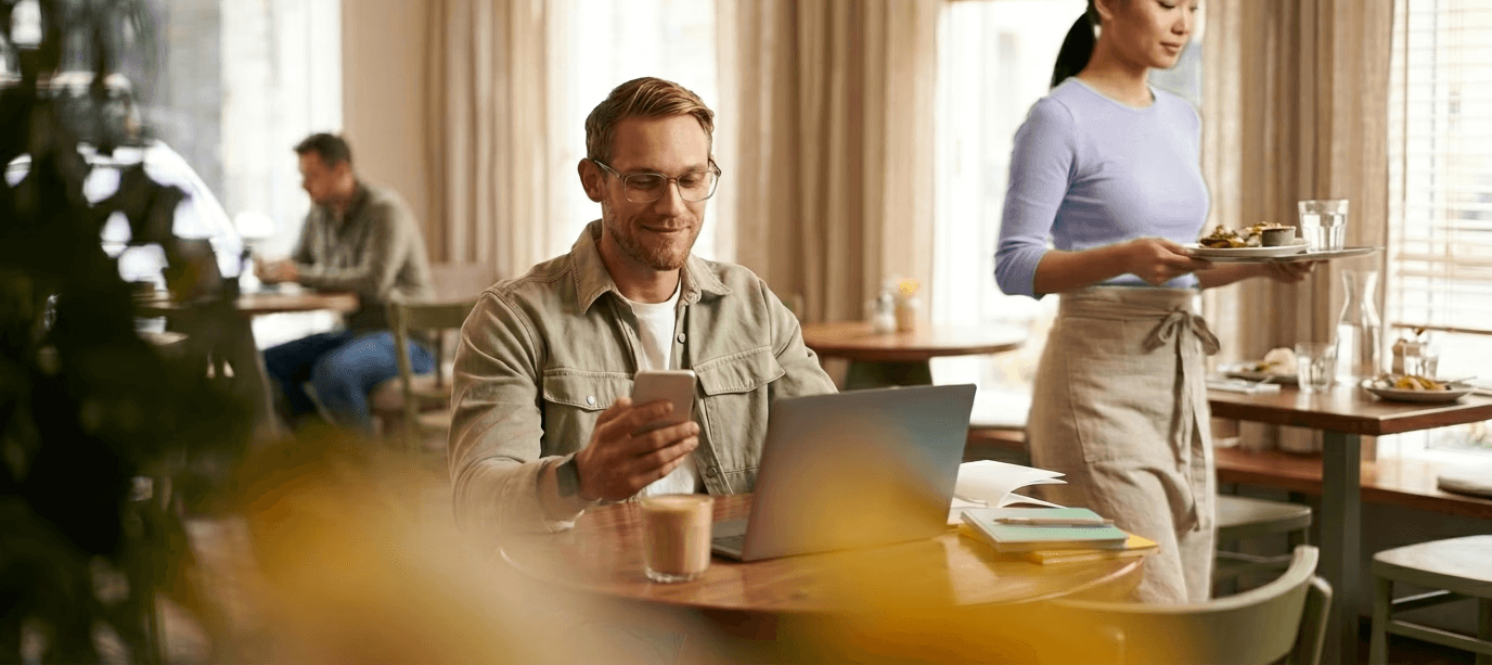 Man using phone and laptop at coffee shop, working remotely
