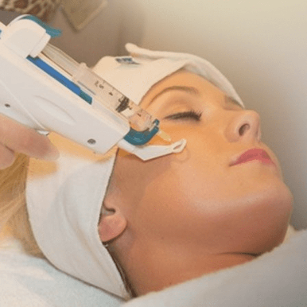 Close-up van een glazen pipet die een enkele druppel helder en rijk serum op de wang van een vrouw laat vallen. De huid ziet eruit als 'glas huid'—extreem dauwachtig en stralend. Zachte warme verlichting. De achtergrond is een romige, off-white waas. Stijl: Hoogwaardige architectonische fotografie met 8k resolutie, zacht daglicht, minimalistische luxe, neutraal kleurenpalet (Beige Taupe Crème Zacht Goud).