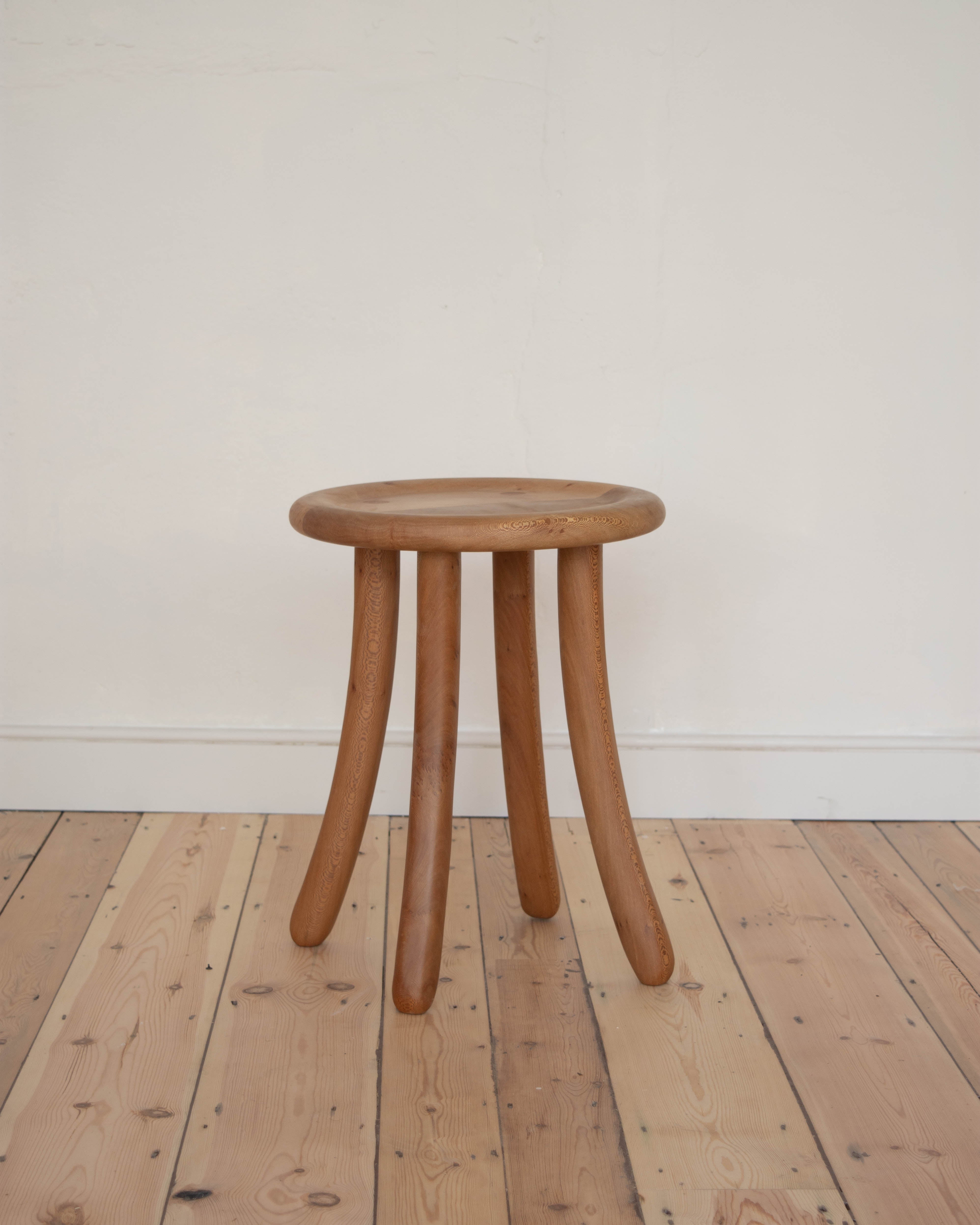 Wooden Luneta stool with four curved legs on a wooden floor against a white wall