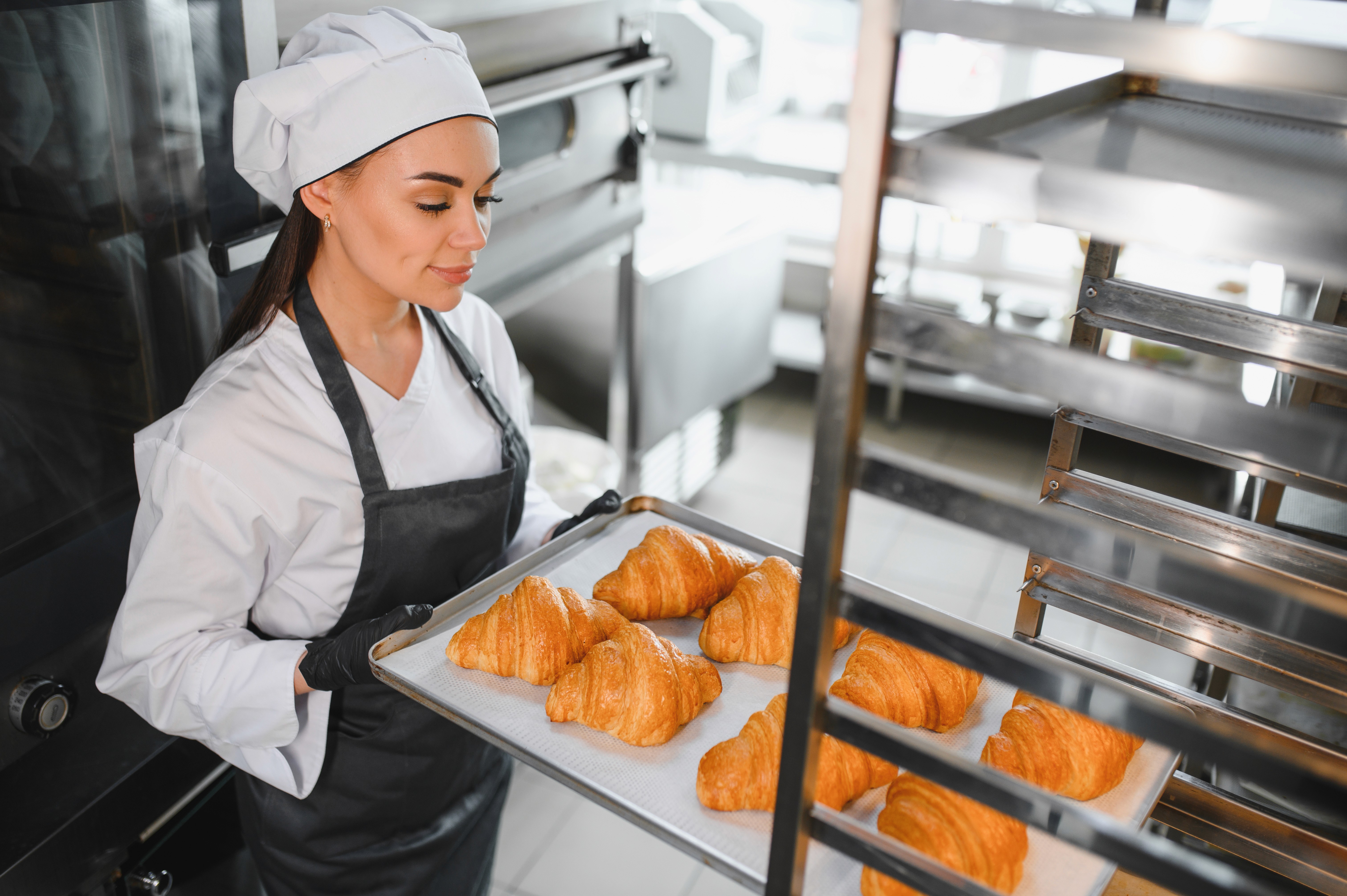 Piekarka wkłada blachę piekarniczą z croissantami do wózka piekarniczego w profesjonalnej piekarni