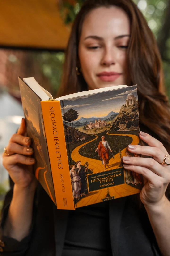 A woman with long hair and a black blazer holds a copy of Boethius's "The Consolation of Philosophy" next to her face, published by Logos.
