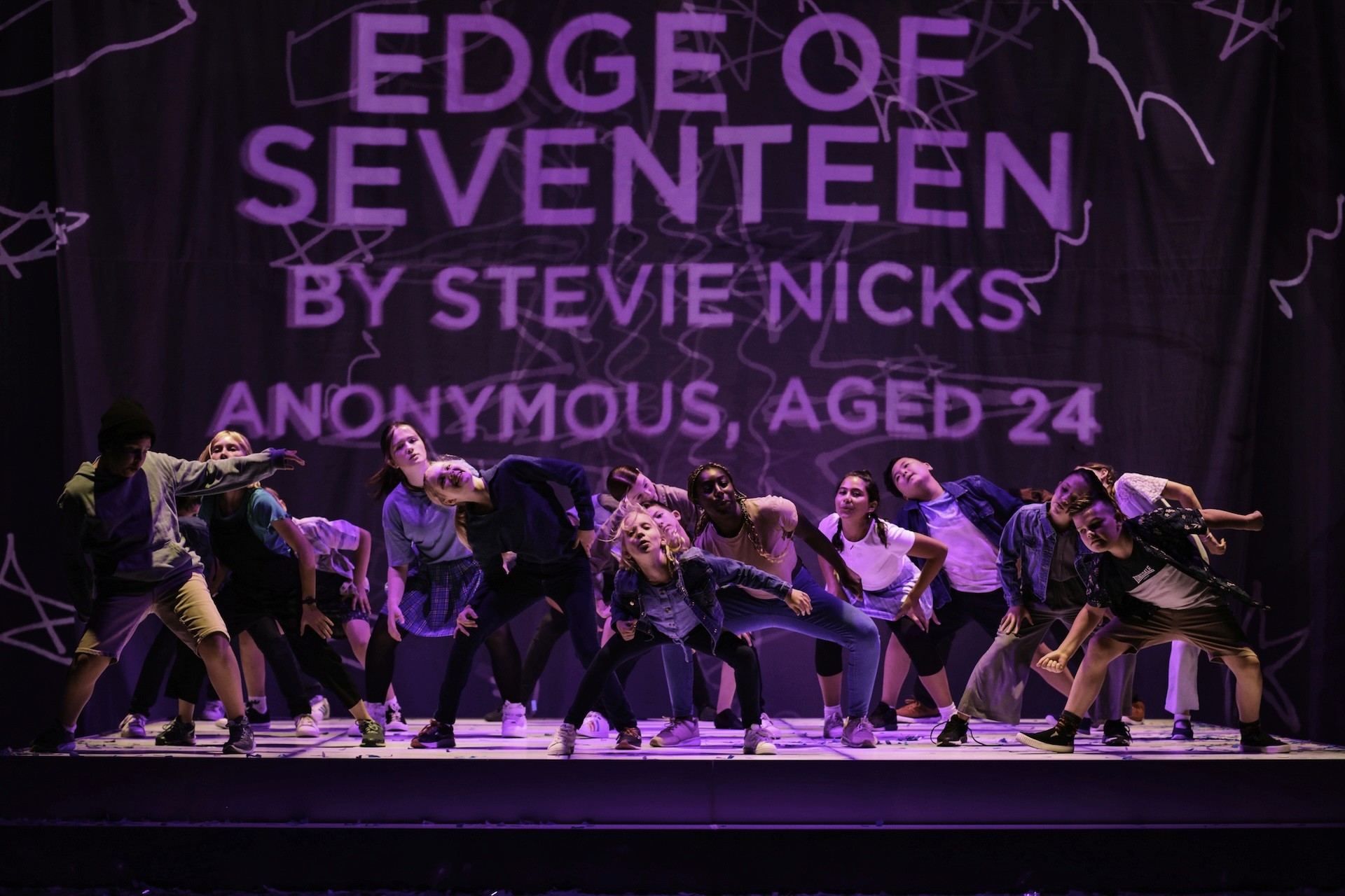 A group of young performers strike a low, energetic dance pose beneath a large projection reading “Edge of Seventeen” by Stevie Nicks. The cast leans forward with playful intensity under purple stage lighting, creating a bold, unified image on a wide theatrical stage.