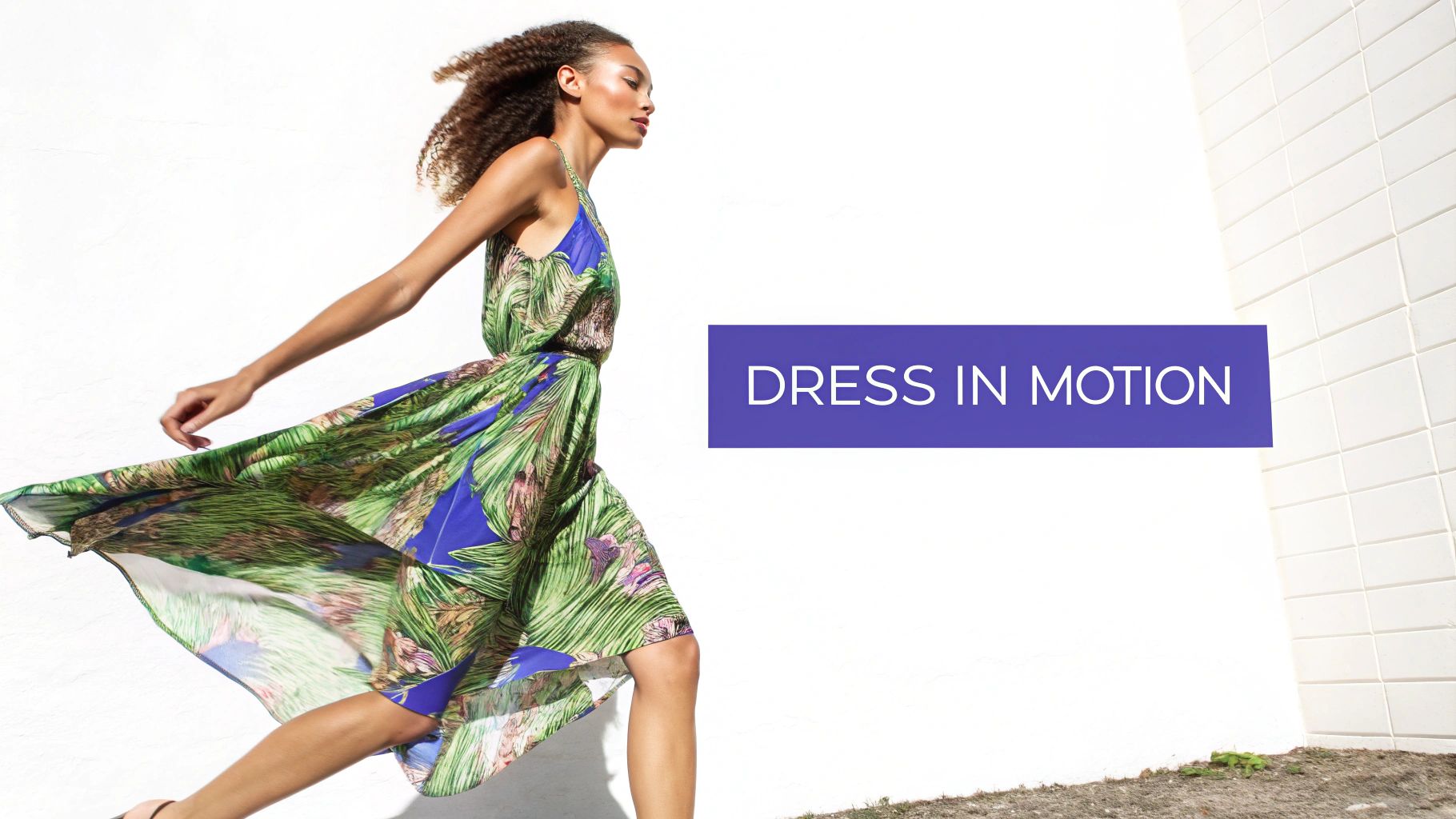 A woman with curly hair in a flowing green and blue tropical dress is in motion against a white wall.