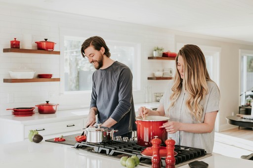 Un homme et une femme cuisinent ensemble dans une cuisine moderne et lumineuse. L'homme coupe des légumes tandis que la femme remue une grande casserole rouge sur une cuisinière. L'image est encadrée avec des coins arrondis.