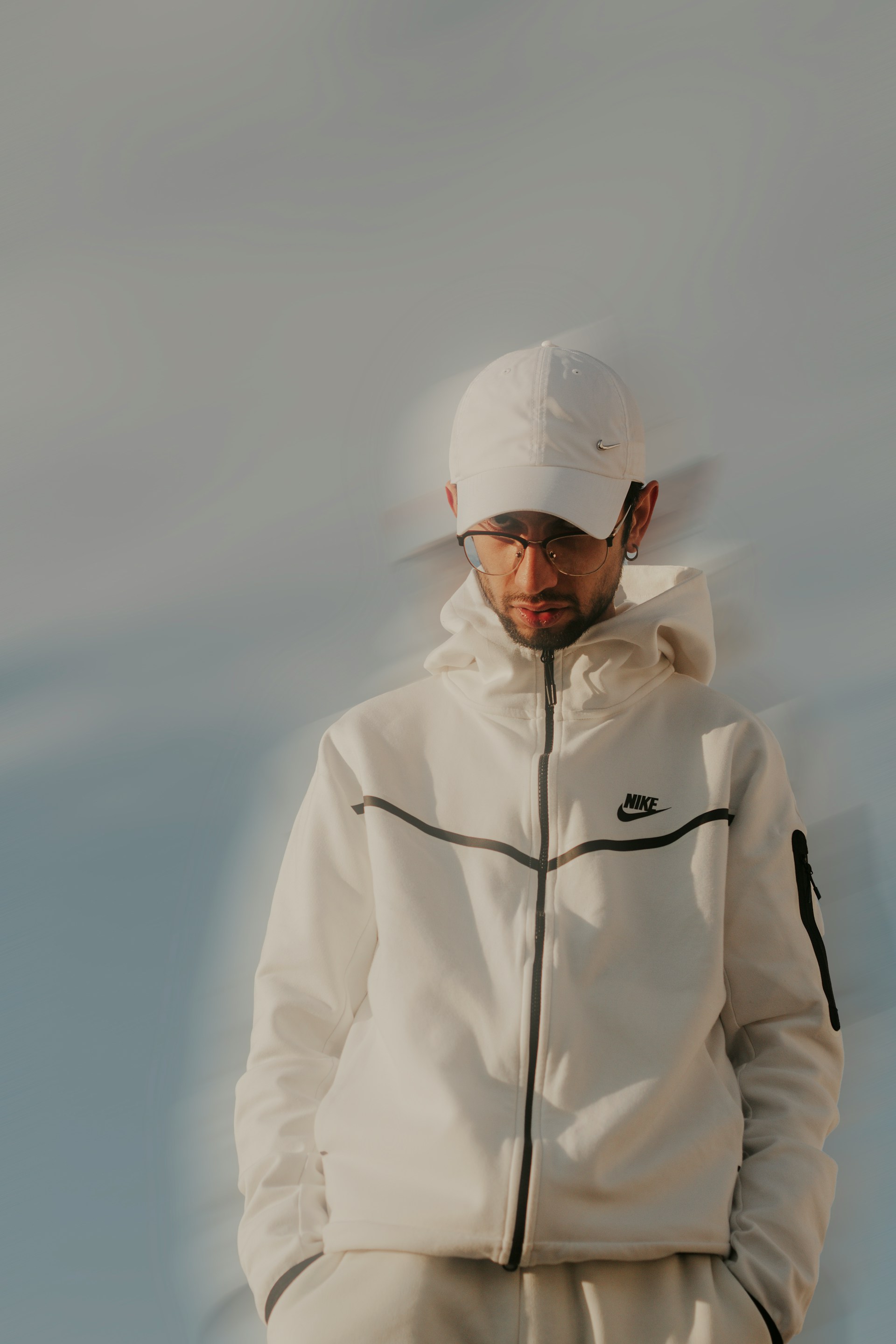 A person wearing a white Nike zip-up hoodie with black detailing and a matching white Nike baseball cap, glasses, and facial hair, posed against a smooth gradient background of light blue and gray tones.
