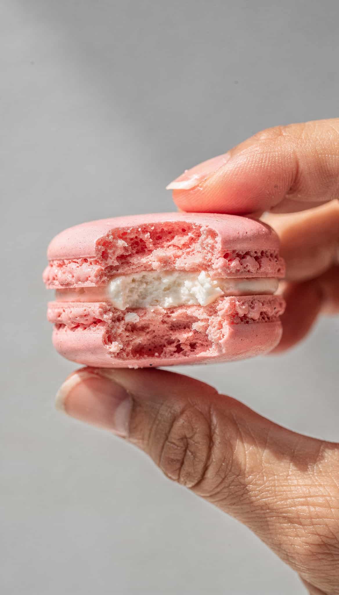 a pink macaron with a bite taken out of it, held between two hands against a plain, light gray background