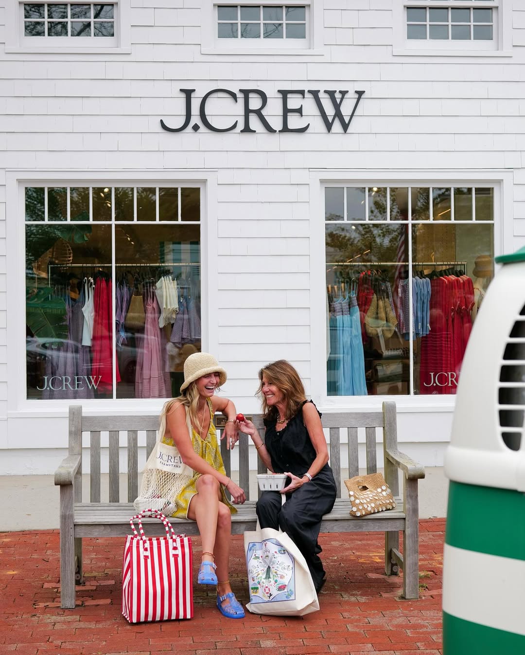 J.Crew storefront sign mounted on a white clapboard façade above large retail display windows, with two shoppers sitting on a bench outside the store.