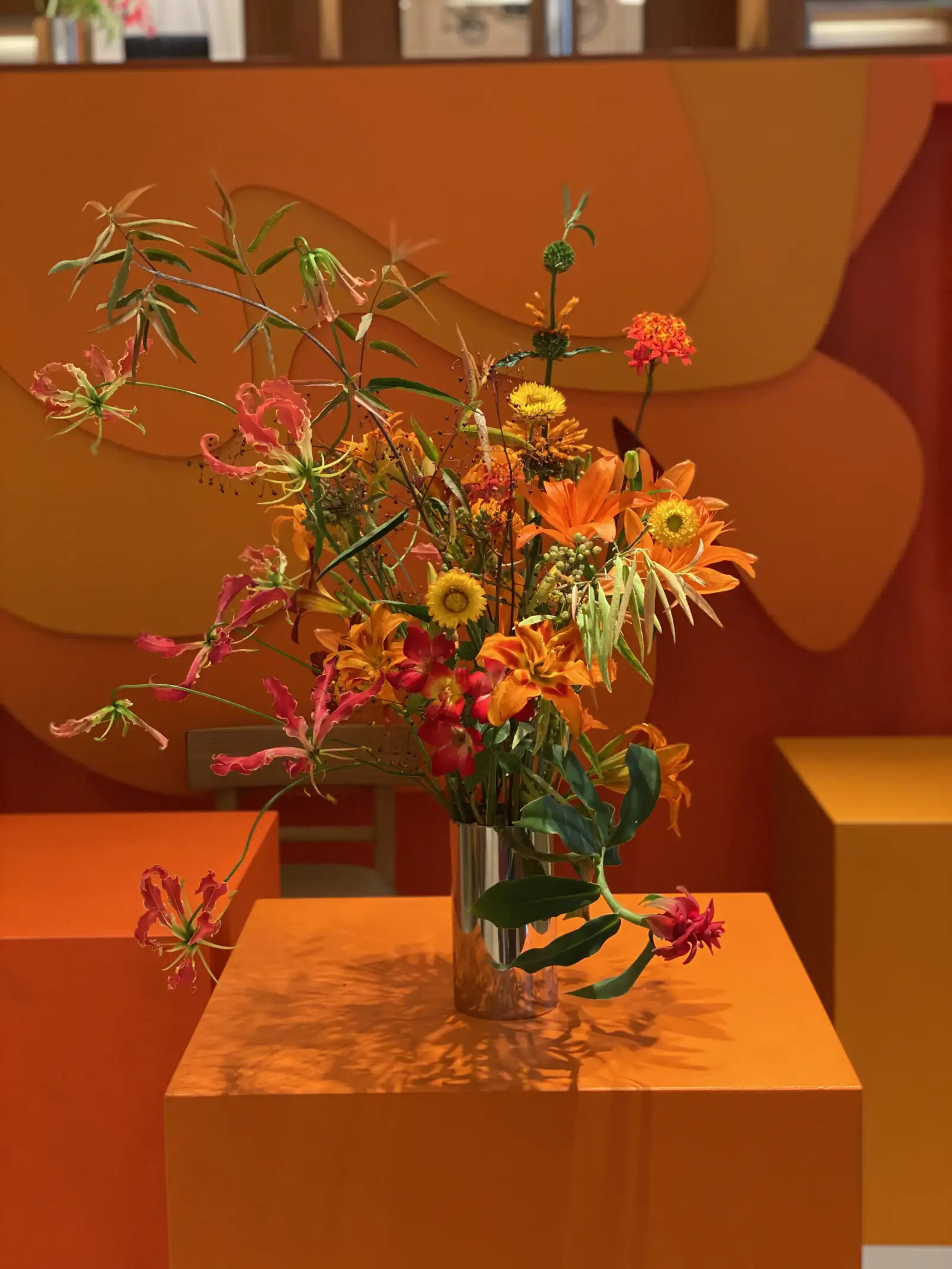 Arreglo floral vibrante en jarrón metálico colocado sobre una base naranja. La composición combina flores exóticas en tonos rojos, naranjas y amarillos con ramas verdes alargadas. El fondo presenta paneles geométricos en degradé cálido, creando una atmósfera moderna y artística con gran armonía cromática.