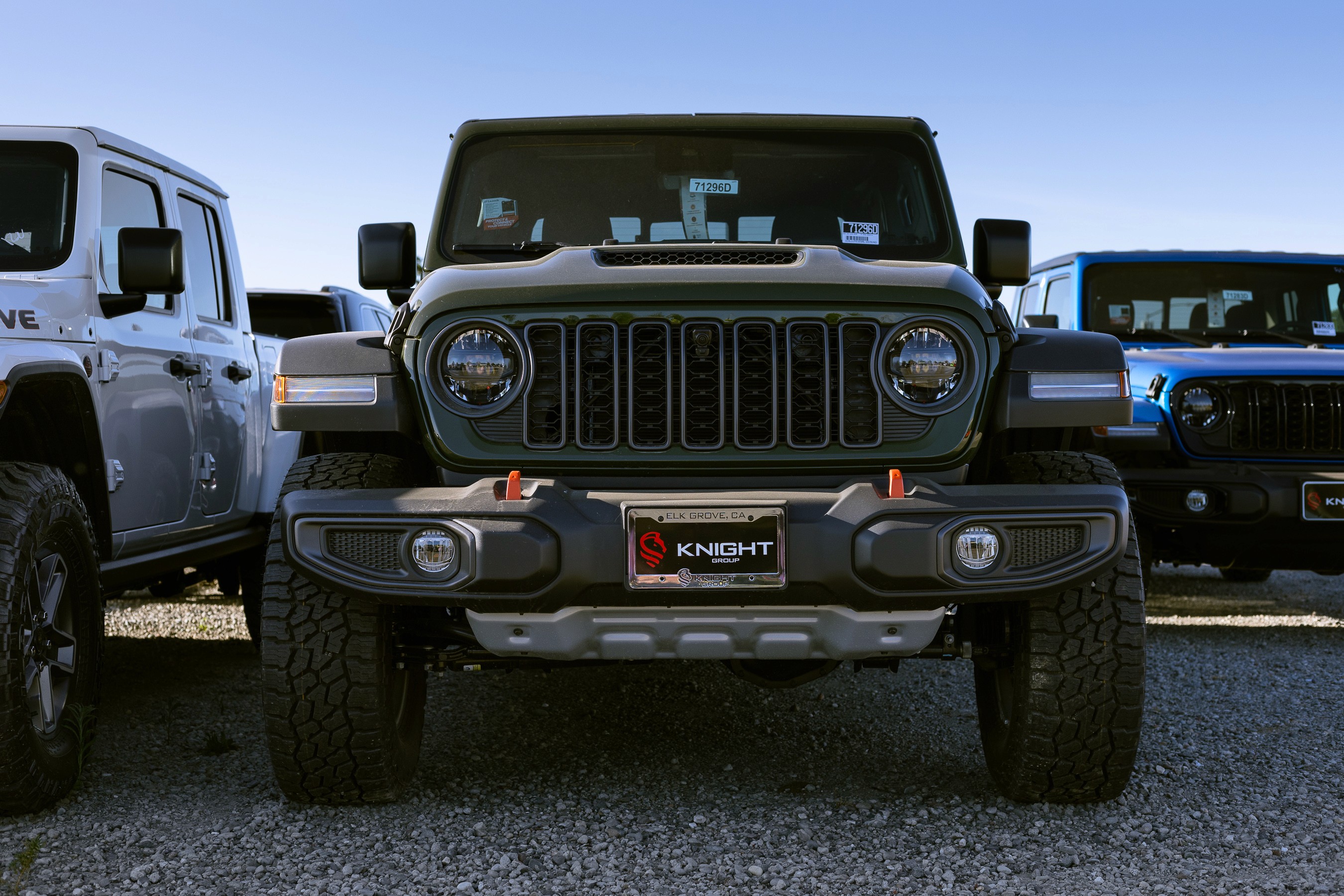 A rugged, green off-road vehicle is parked on gravel, flanked by other vehicles, featuring a prominent front grille and large tires under a clear blue sky.