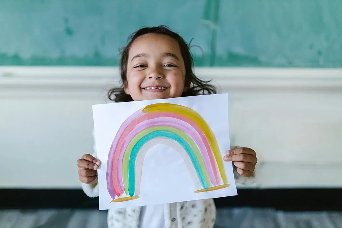 Child holds up rainbow drawing, smiling.