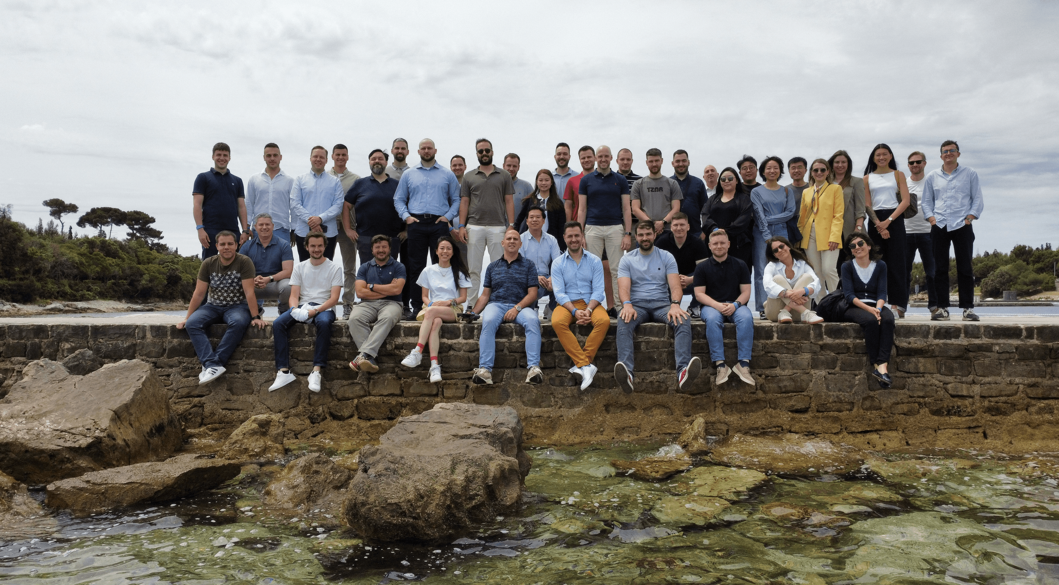 A large group of people standing together on a rocky shoreline, with a cloudy sky in the background.