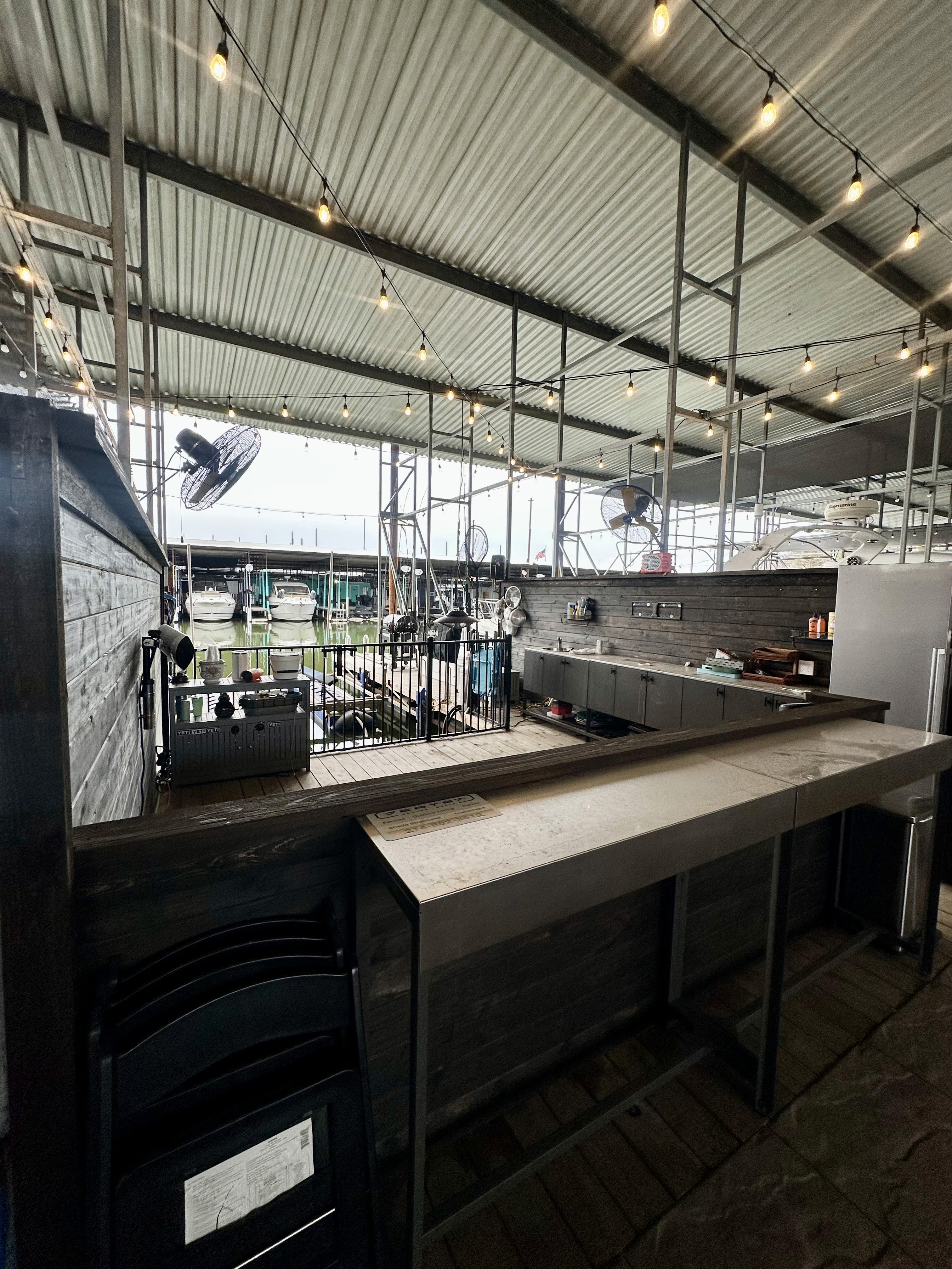 A modern industrial-style outdoor kitchen area features stainless steel appliances, string lights, and a corrugated metal roof, creating a sleek and functional cooking space with wooden and metal accents.