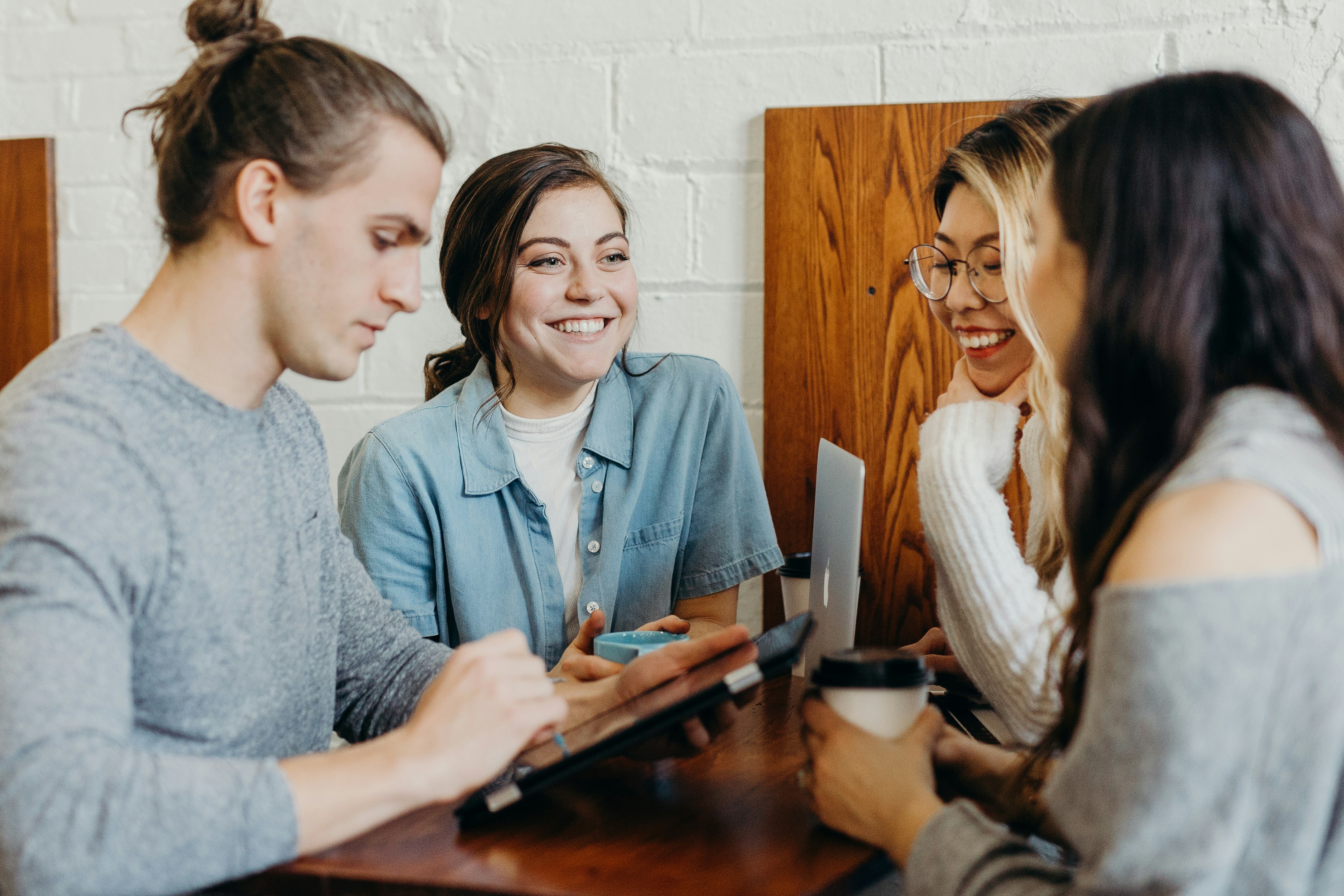 Four young adults gathered around a small table in a café, engaging in conversation with coffee cups and a digital tablet visible.