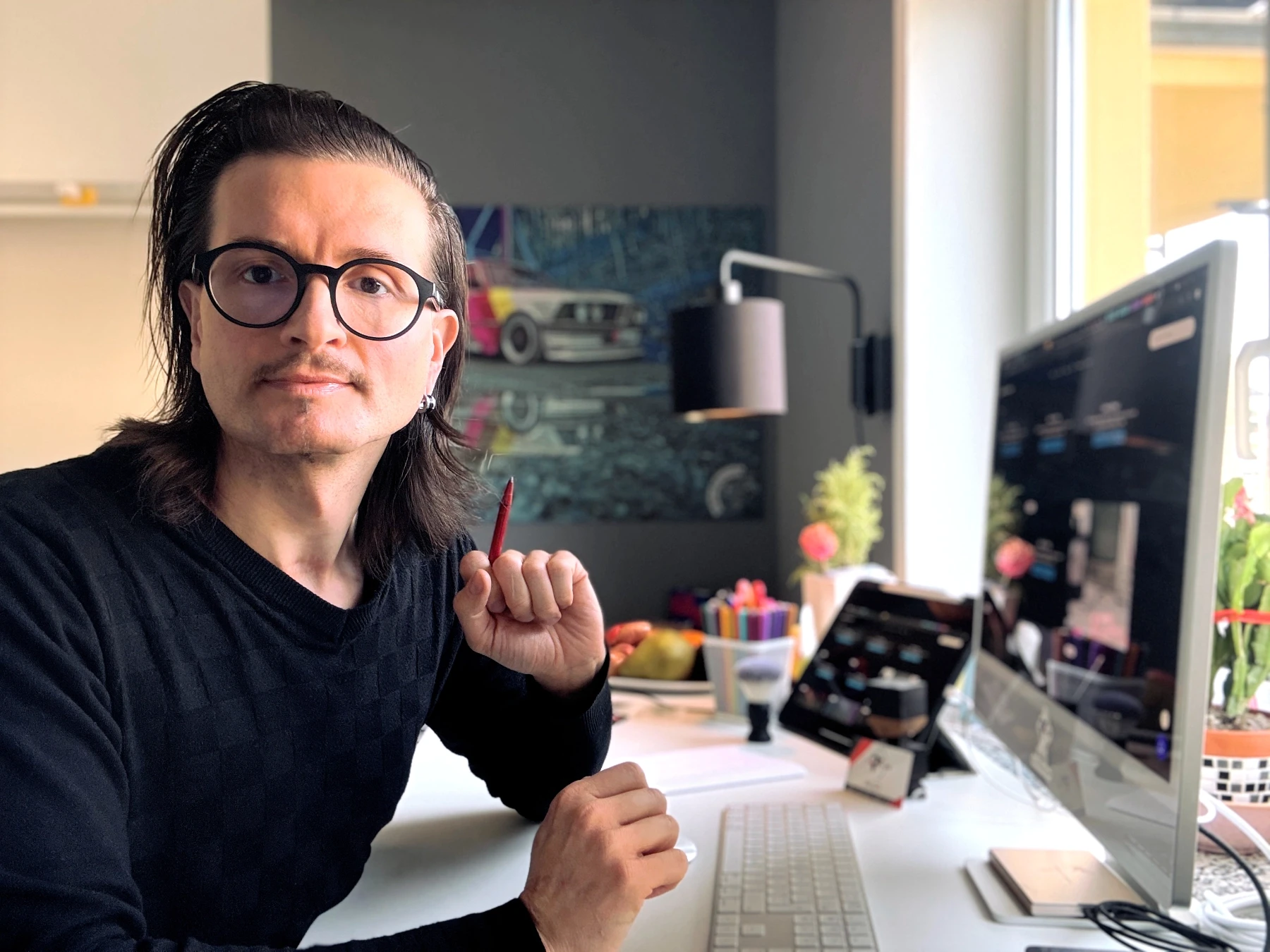 Jens Pfeifer sitzt an seinem aufgeräumten Work Desk und blickt konzentriert in die Kamera. Vor ihm stehen ein großer Desktop-Monitor und ein Tablet mit Design-Interfaces, umgeben von Zeichenwerkzeugen, Pflanzen und Studio-Elementen – Sinnbild für strukturierte, kreative und systemische Designarbeit.