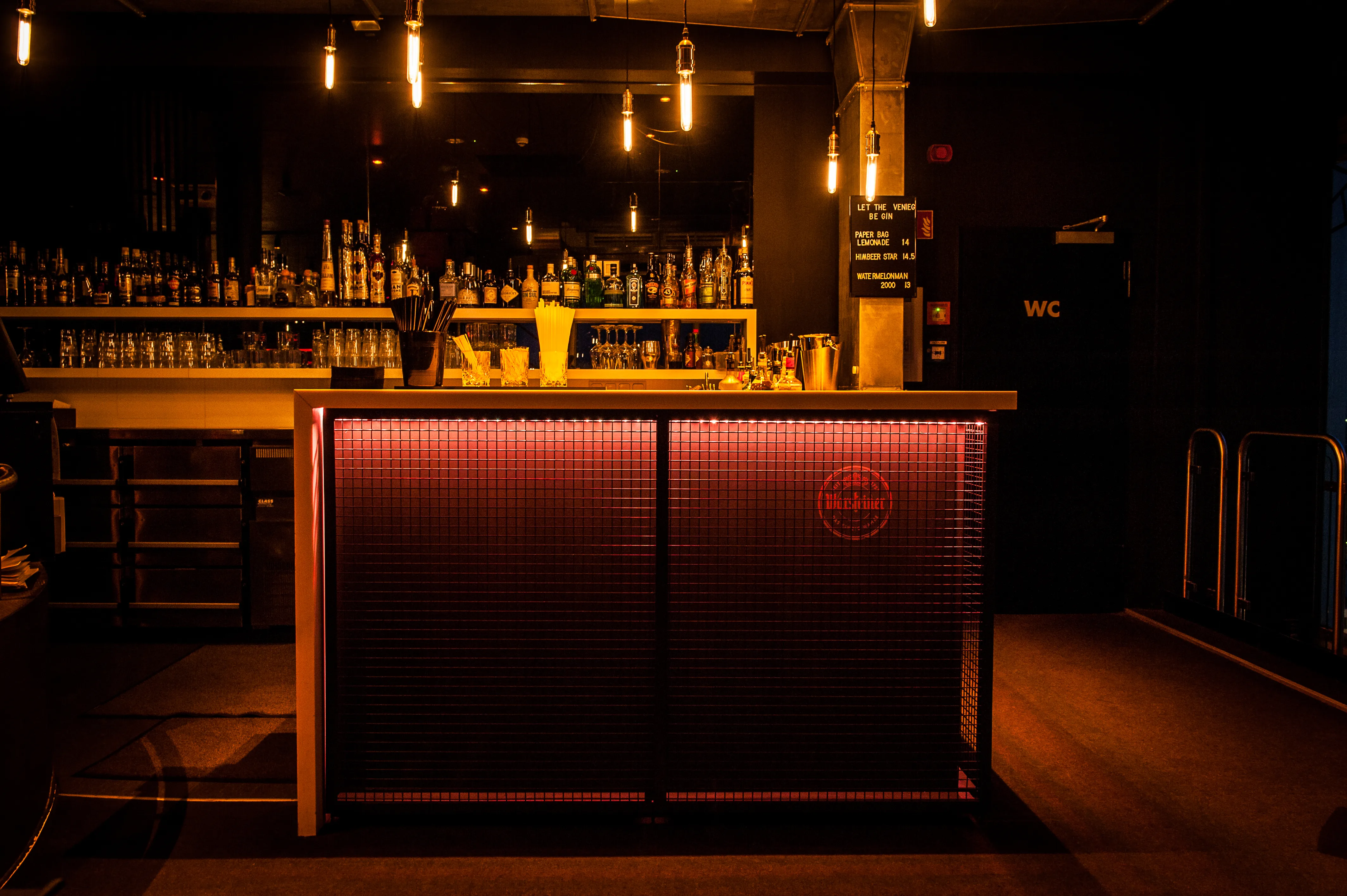 Bar counter at SOLAR SKY LOUNGE clad in red-lit metal mesh with a Warsteiner logo, backed by shelves of spirits and glowing filament pendant bulbs.