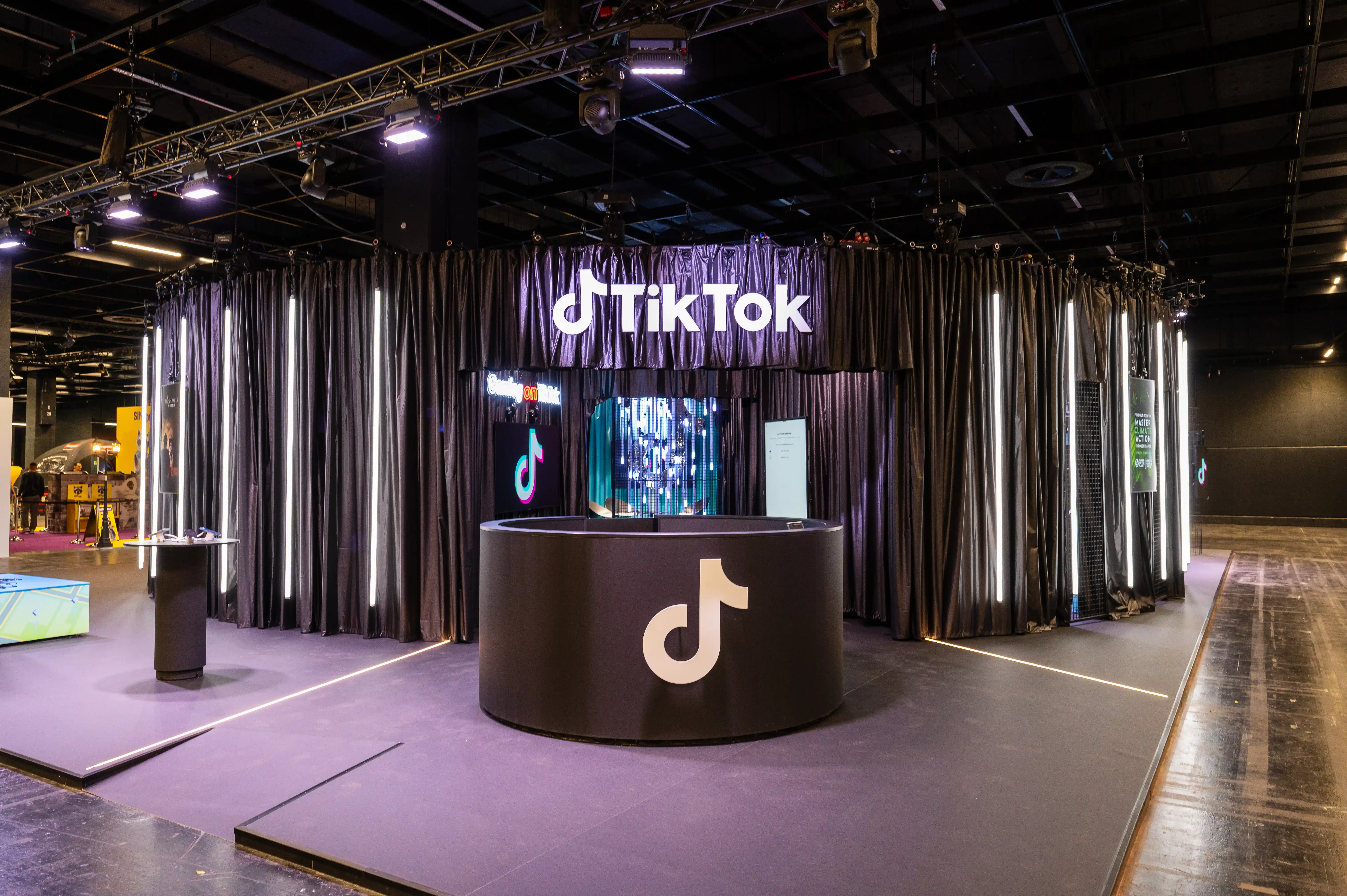 TikTok booth at Gamescom with round logo counter in front of black curtains, LED screen and glowing lettering.