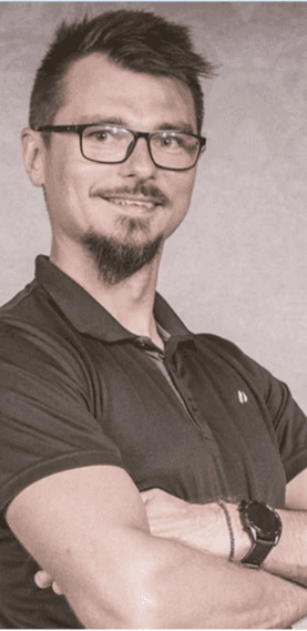 Zdjęcie przedstawia kobietę o jasnych, falowanych włosach i okularach w ciemnych oprawkach. Ma na sobie czarną bluzkę z wzorem i patrzy prosto w obiektyw. Tło jest jednolite, ciemne.