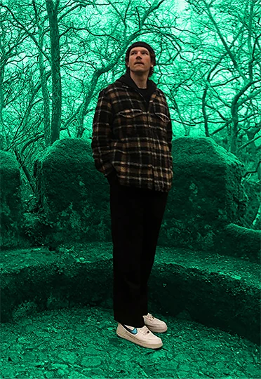 Ein Mann steht in einer surreal wirkenden, grün getönten Waldlandschaft mit Moos bedeckten Steinen und Bäumen. Er trägt eine Mütze, ein kariertes Hemd, schwarze Hosen und helle Sneaker. Sein Blick ist nach oben gerichtet.
