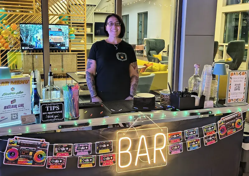 A smiling person stands behind an outdoor bar setup in Bradenton, FL, with a variety of liquor bottles and cocktail-making tools arranged neatly, set against the backdrop of a serene lake and lush greenery.