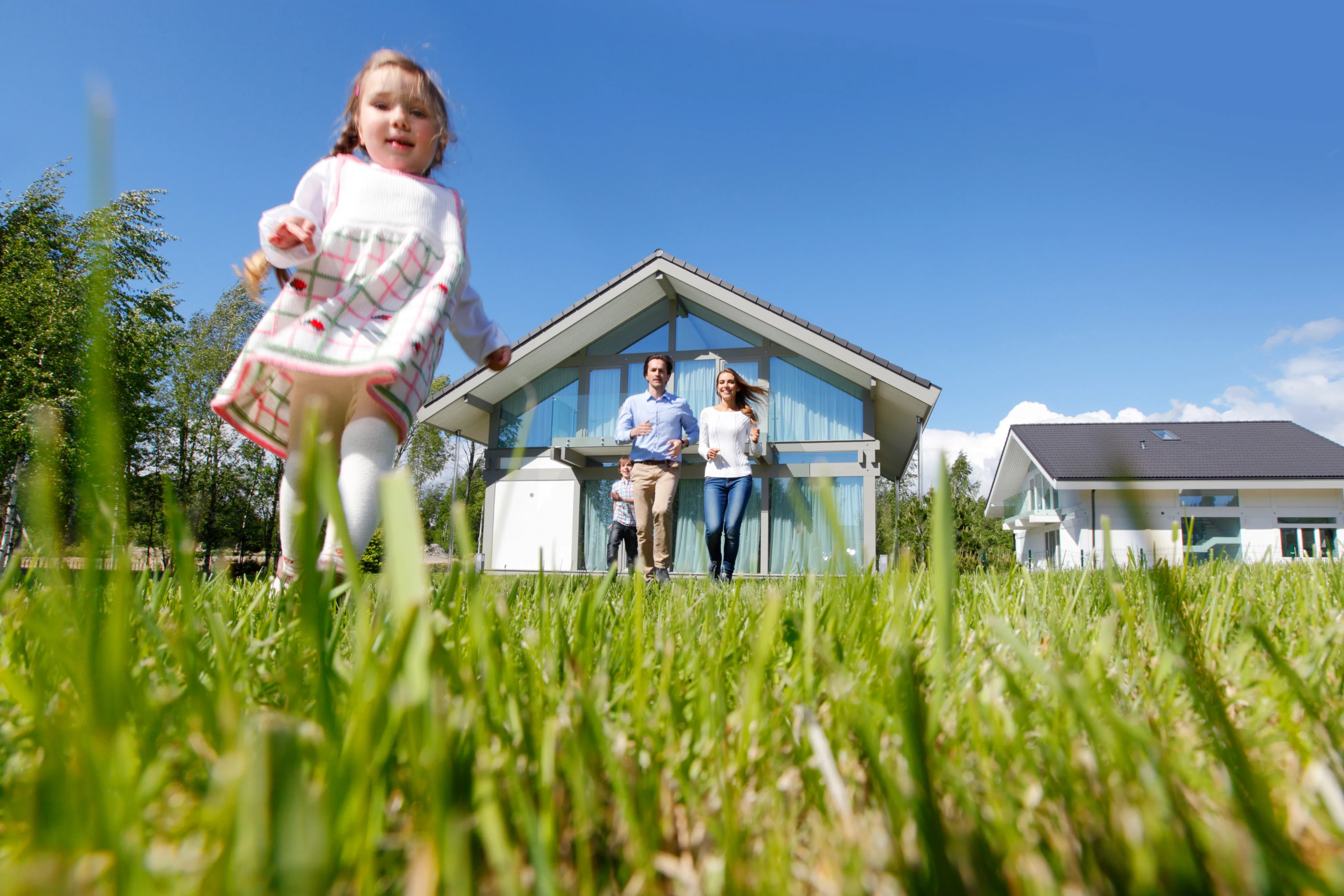 A young girl in a pink and white outfit runs through a grassy lawn toward her family standing in front of a modern white house on a sunny day.