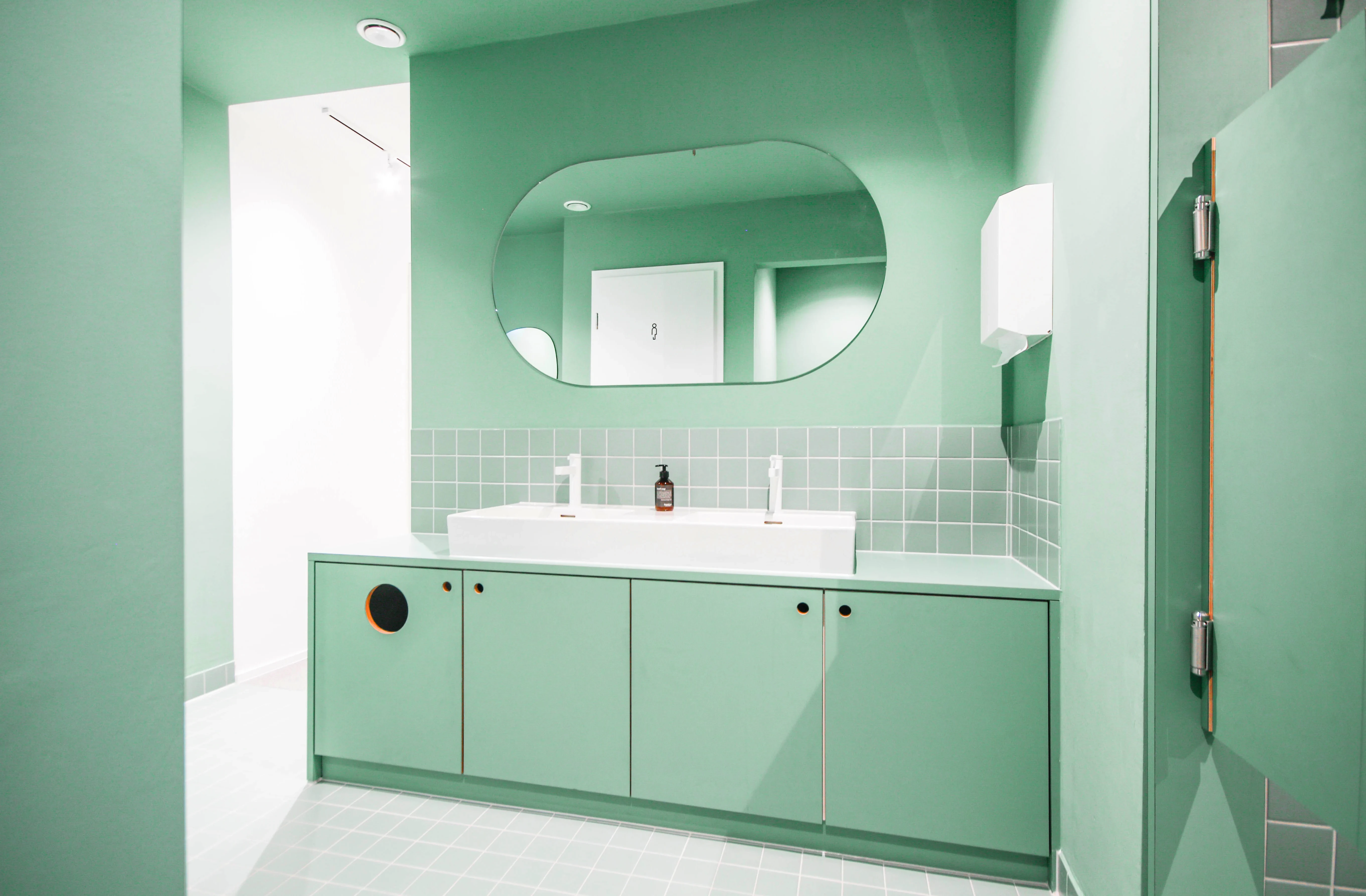 Mint-green bathroom at CYCLA with a long vanity unit, white double washbasin, oval mirror on a green wall and matching green shower doors.