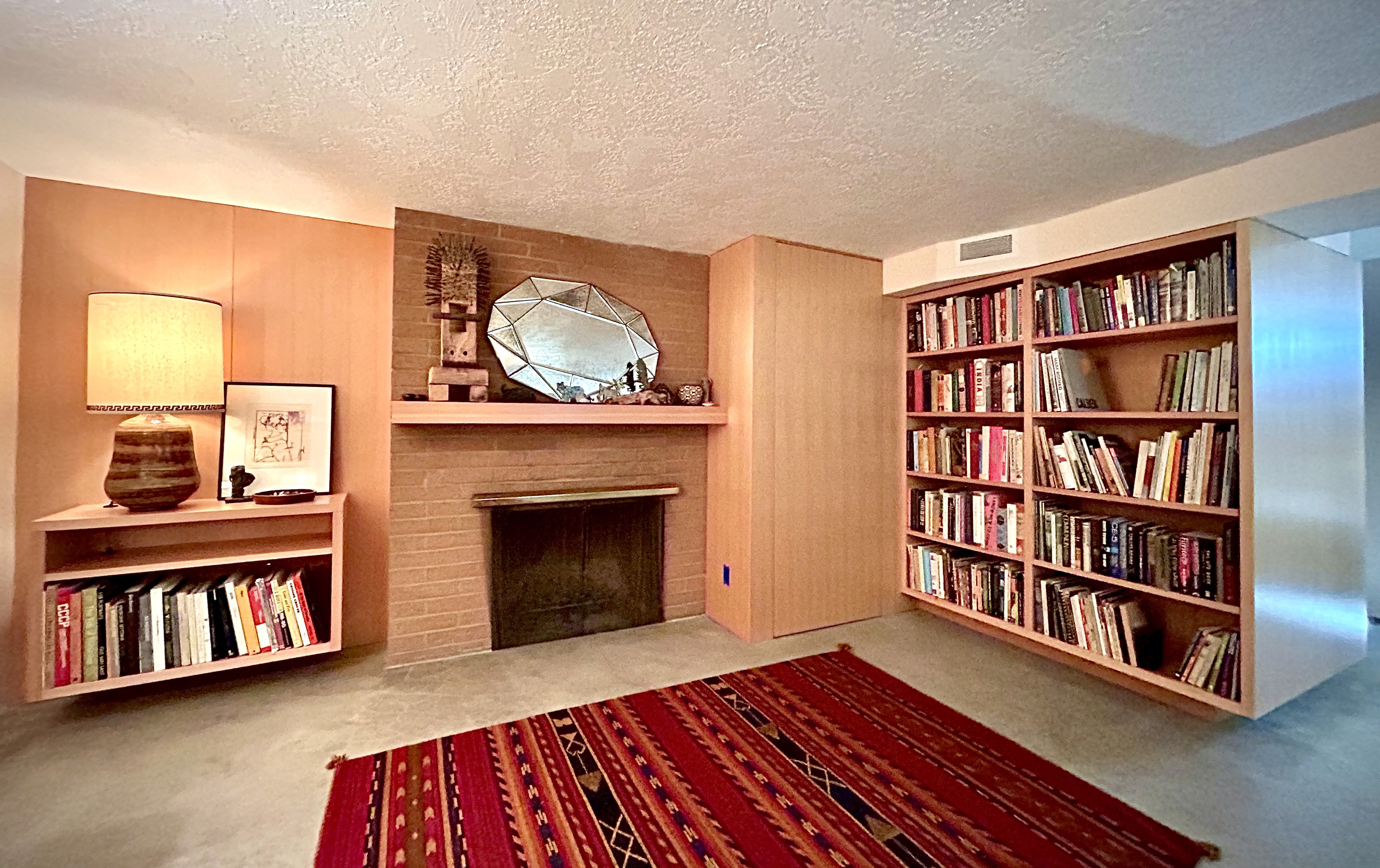 A view of a den with fireplace, wooden mantel and wall panel, floating cabinet, and double bookshelf with a colorful patterned carpet in the foreground.