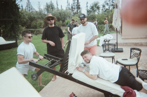 polaroid photo of group of people resting and having fun together. Some of people are looking to the camera. The man on the right side of the pic is laying on the sun lounger, the other man on the left side took the lounger and picked it up for fun