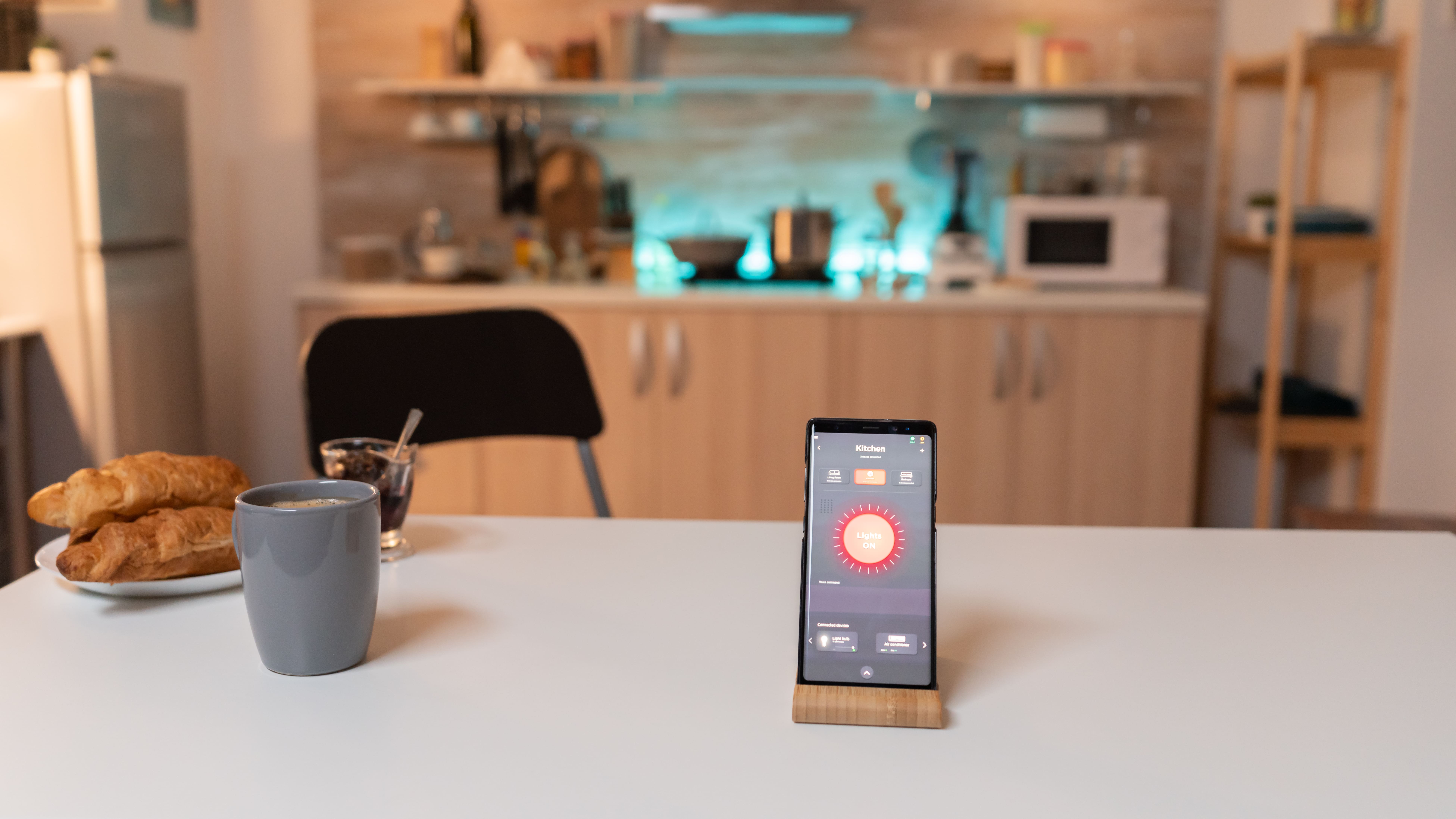 A smartphone displaying a smart home app for controlling kitchen lights sits on a breakfast table with coffee and croissants, with the modern, automated kitchen blurred in the background.