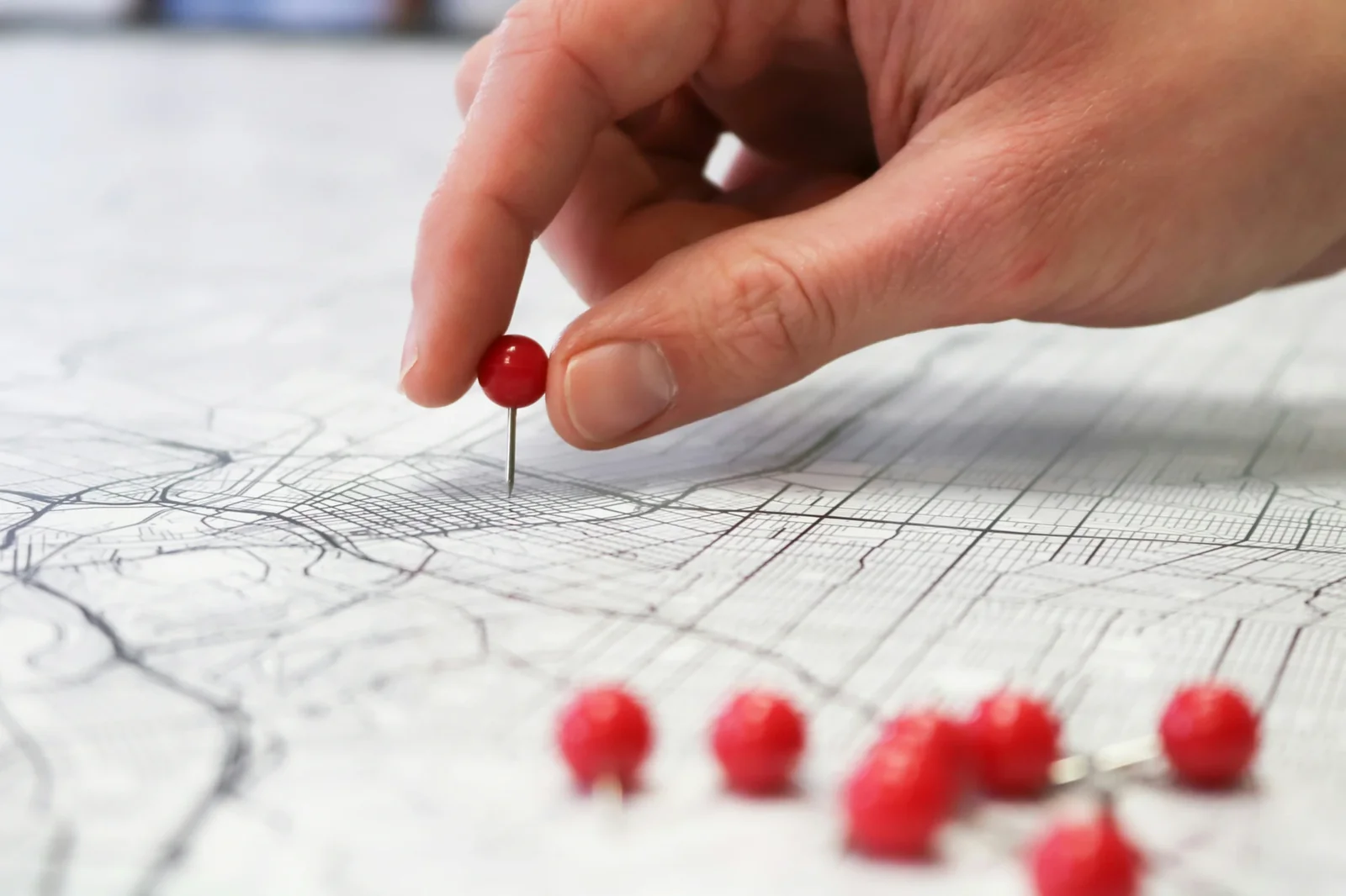 Hand placing red pushpin on map with scattered pins marking geographic locations.