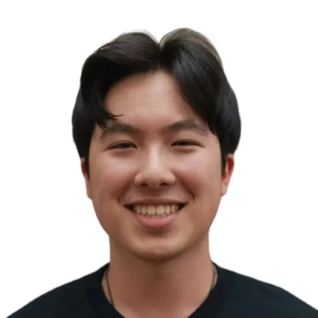 A young man with dark hair smiles warmly against a plain white background, wearing a black shirt.