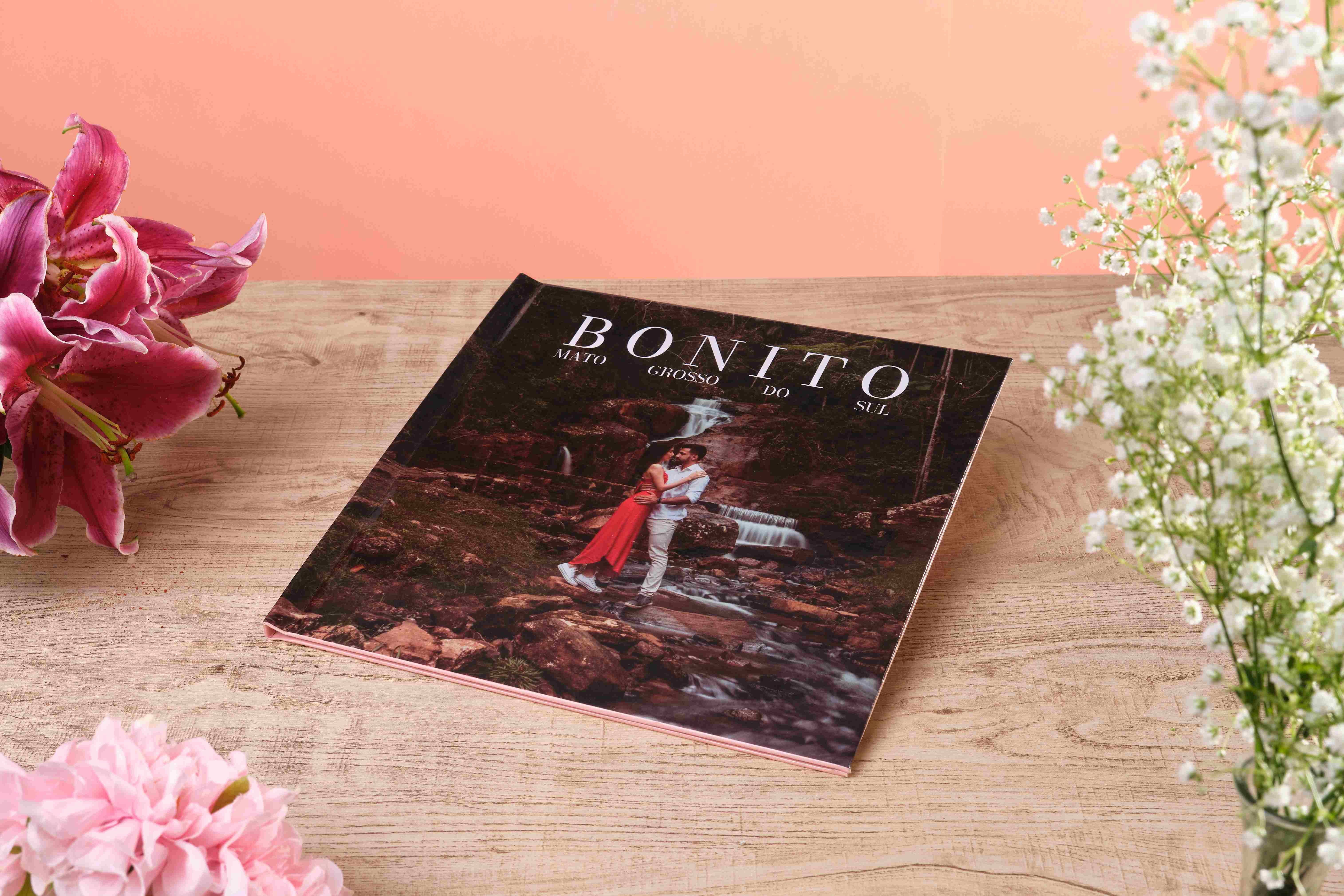 Mãos segurando fotolivro com capa de Arraial do Cabo sobre mesa de madeira clara, ao lado de flores cor-de-rosa e lilás