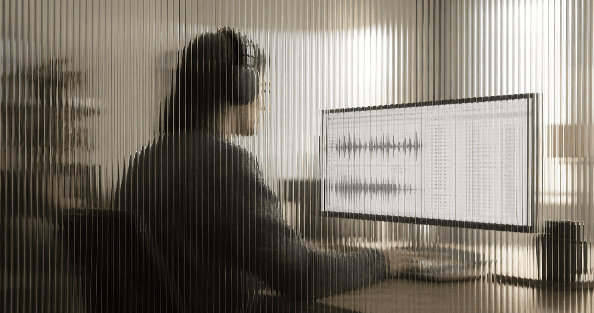 Analyst with headphones reviewing call recordings and transcripts on a monitor, seen through vertical fluted glass