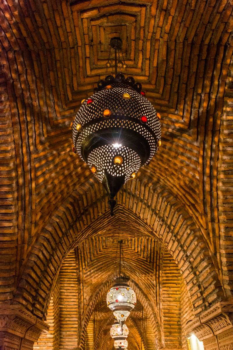 A large, ornate crystal chandelier hangs from a vaulted brick ceiling in a dimly lit, arched hallway with warm lighting.