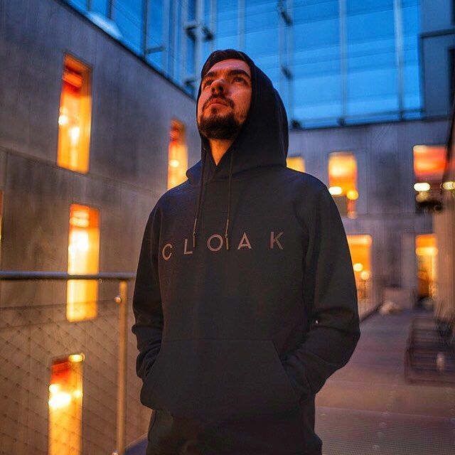 A person wearing a hooded sweatshirt stands in a well-lit space with large windows, looking up thoughtfully.
