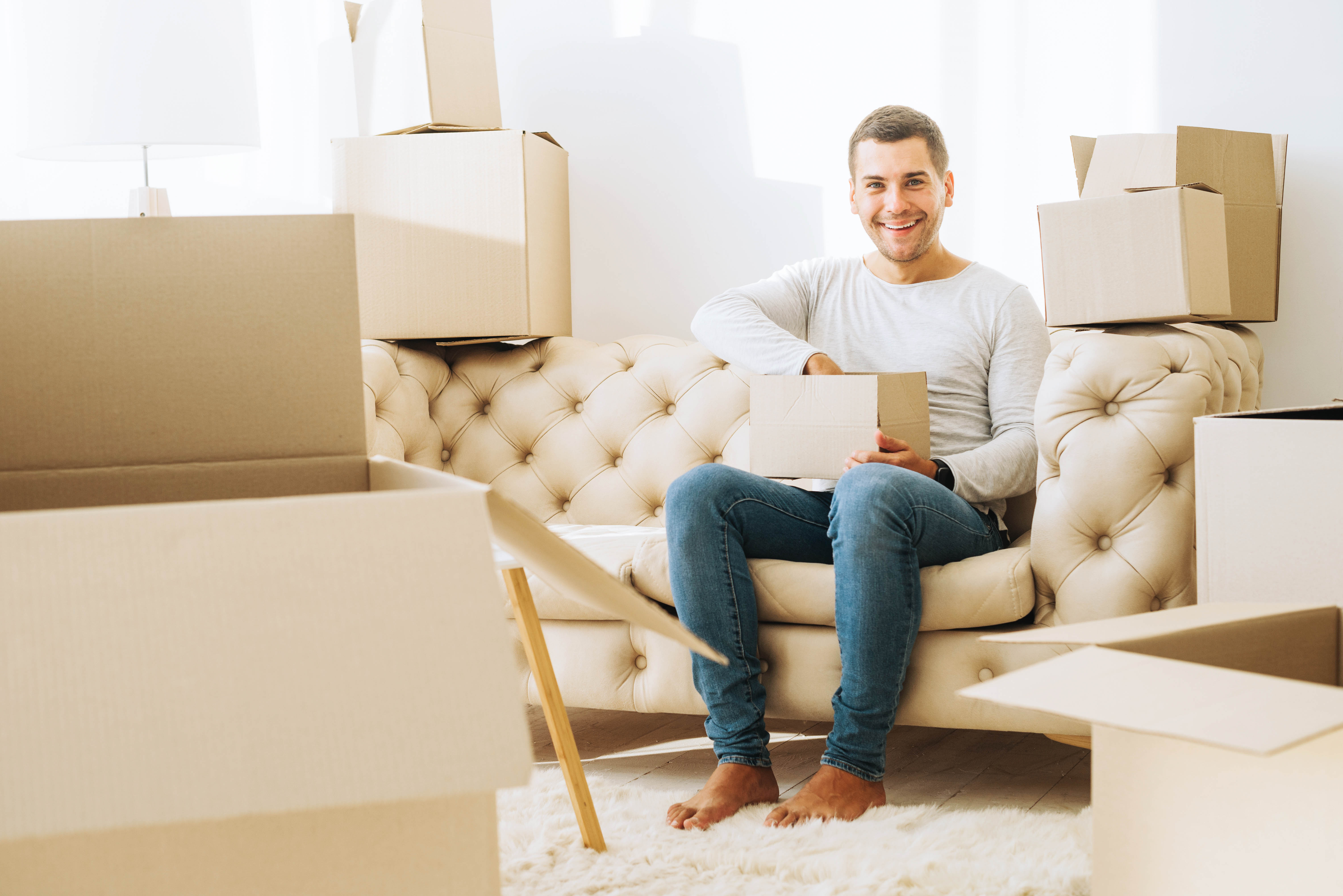 Smiling man sitting on a tufted sofa surrounded by open cardboard boxes during a home relocation process.